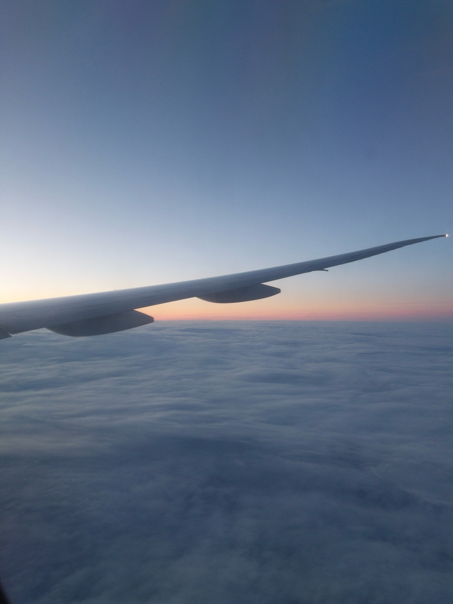 A view of an airplane wing from an airplane window, flying over a cloud cover sky below with the first light of dawn on the horizon.