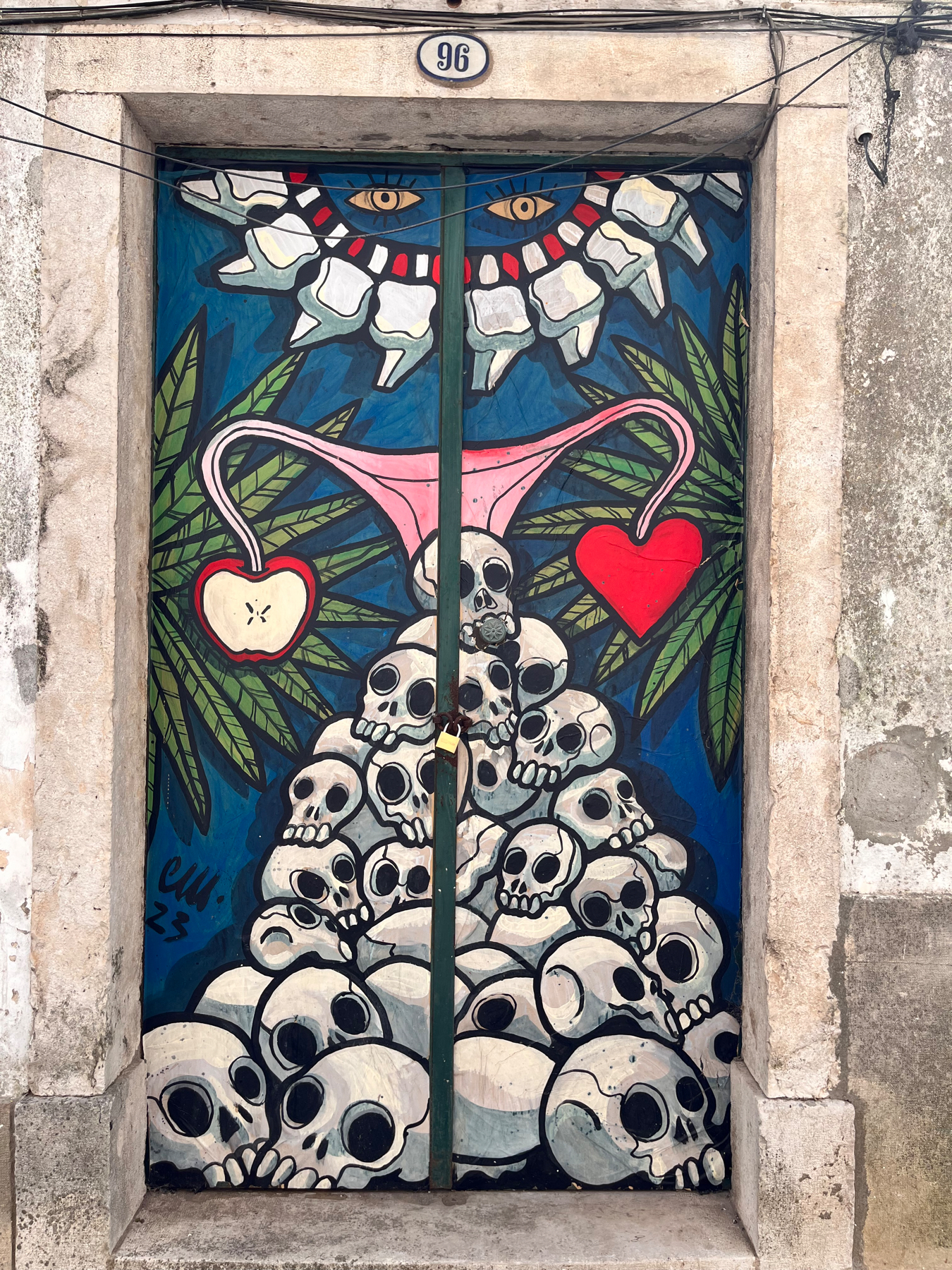 A colorful mural painted on a door depicting a tower of stylized skulls with a background of leaves and two large sets of eyes above. A pink uterus-like shape with a red heart hangs in the center between the eyes, and cut apples on either side