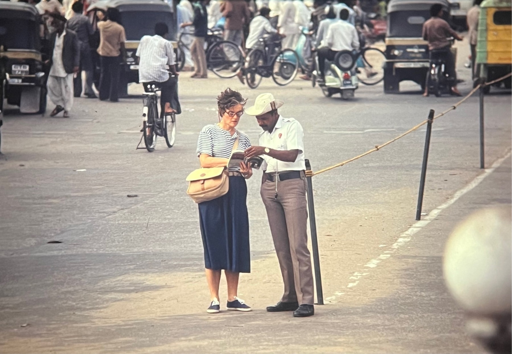 A female tourist and a policeman consulting a book, possibly a travel book, in the middle of a busy Indian road.