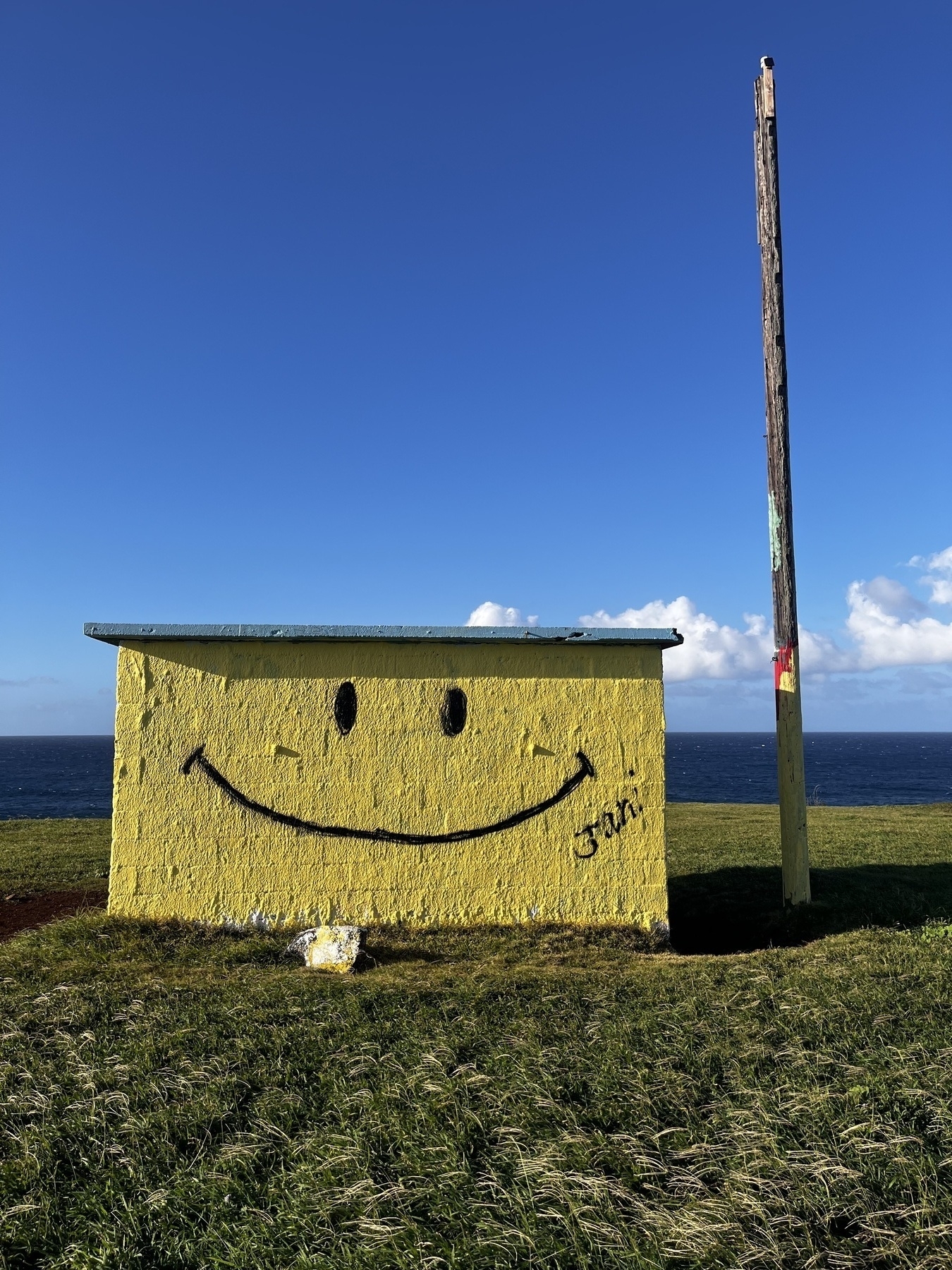 Auto-generated description: A small yellow building by the sea features a large painted smiley face with a wooden pole nearby.