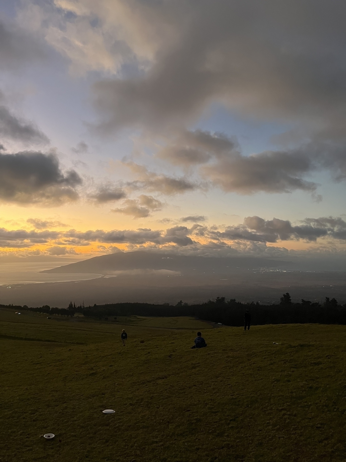 A scenic sunset over a hilly landscape with scattered clouds and distant mountains, accompanied by people relaxing on the grass. The ocean can be seen in the distance.