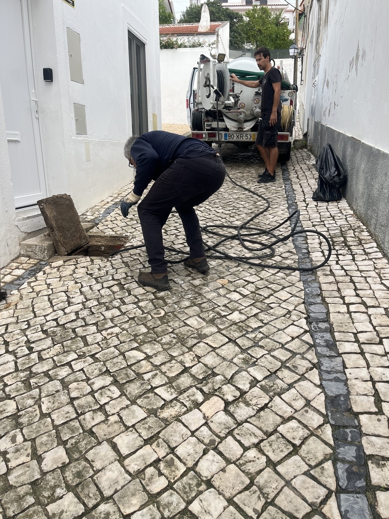 Auto-generated description: Two people are working on a cobblestone street, with one handling a hose near an open manhole and a truck in the background.