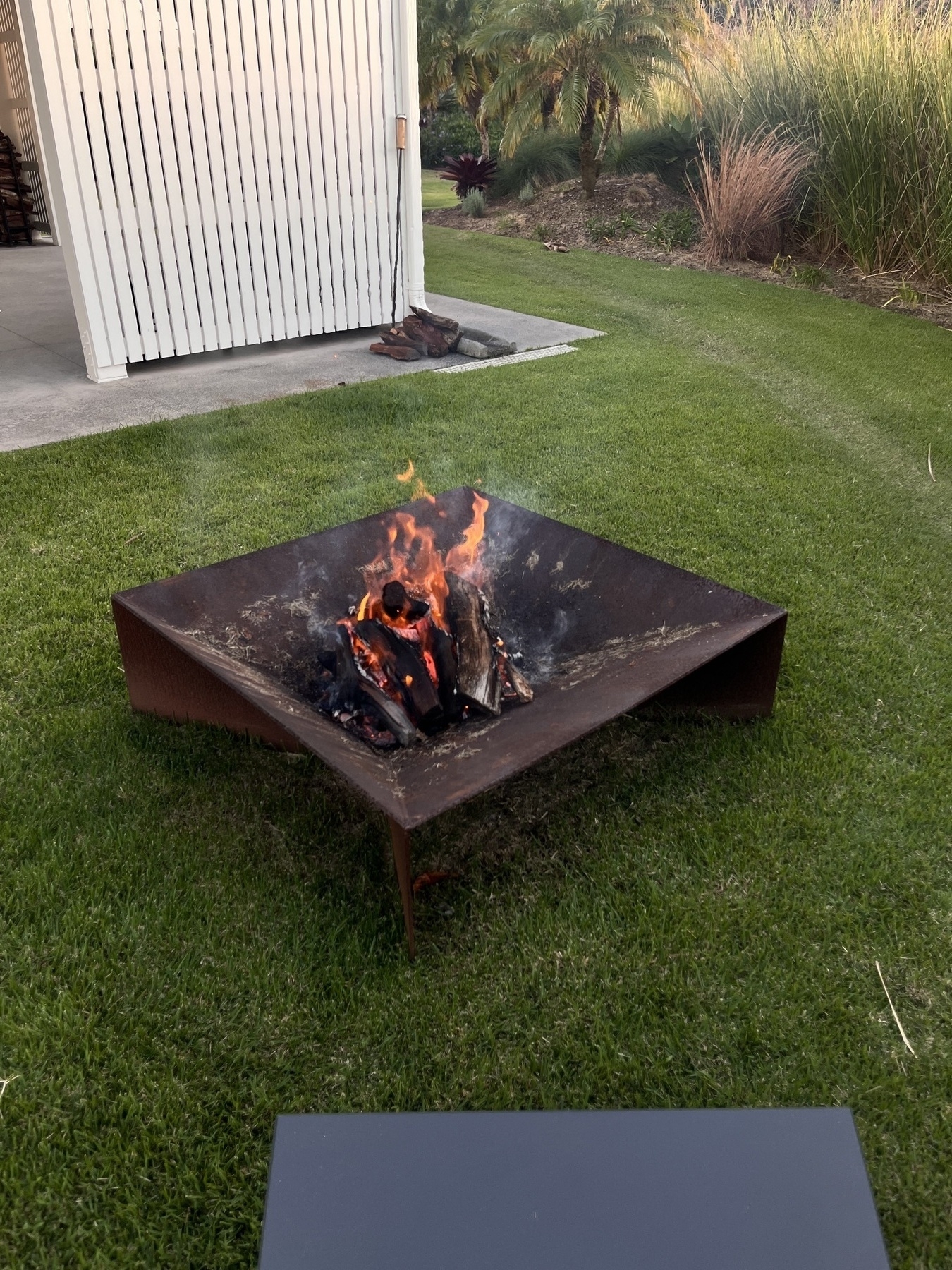 A square metal fire pit with burning logs is set on a grassy lawn near a building.