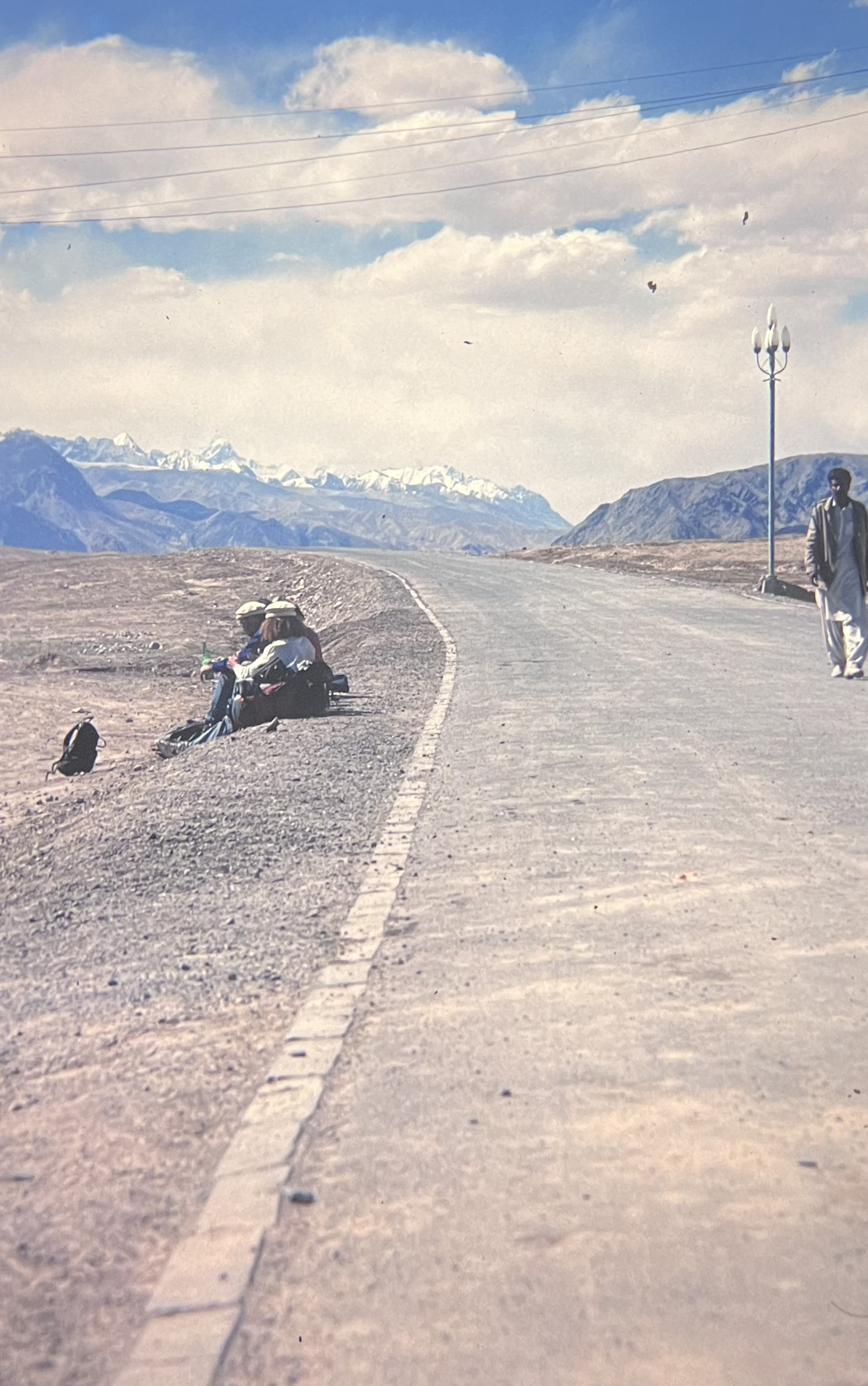 A group of people and their belongings are sitting at the side of a deserted road, with mountainous terrain visible in the background, while another person walks on the opposite side of the road.