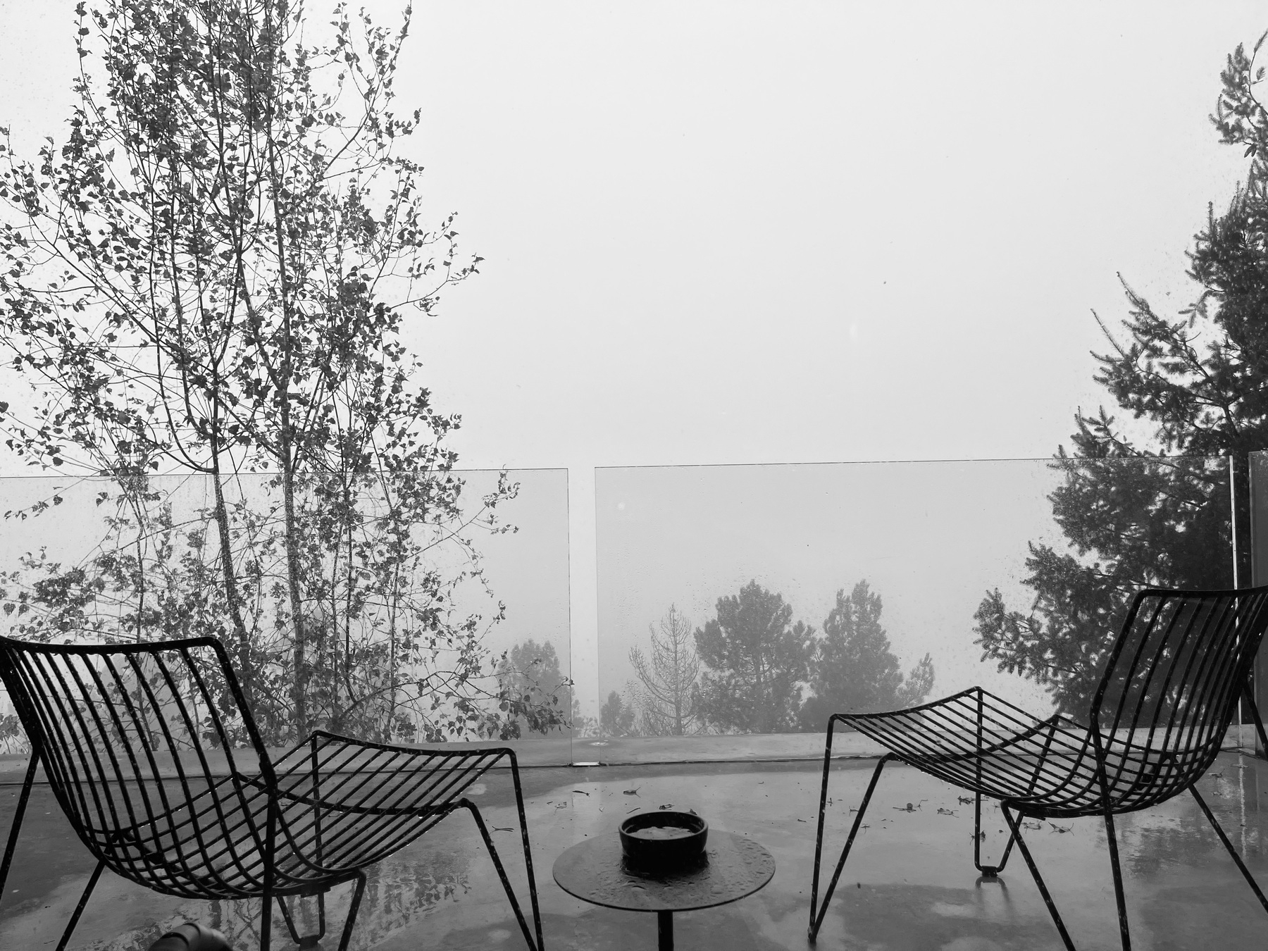 Two empty modern chairs and a small table are positioned on a terrace overlooking a foggy landscape surrounded by trees.