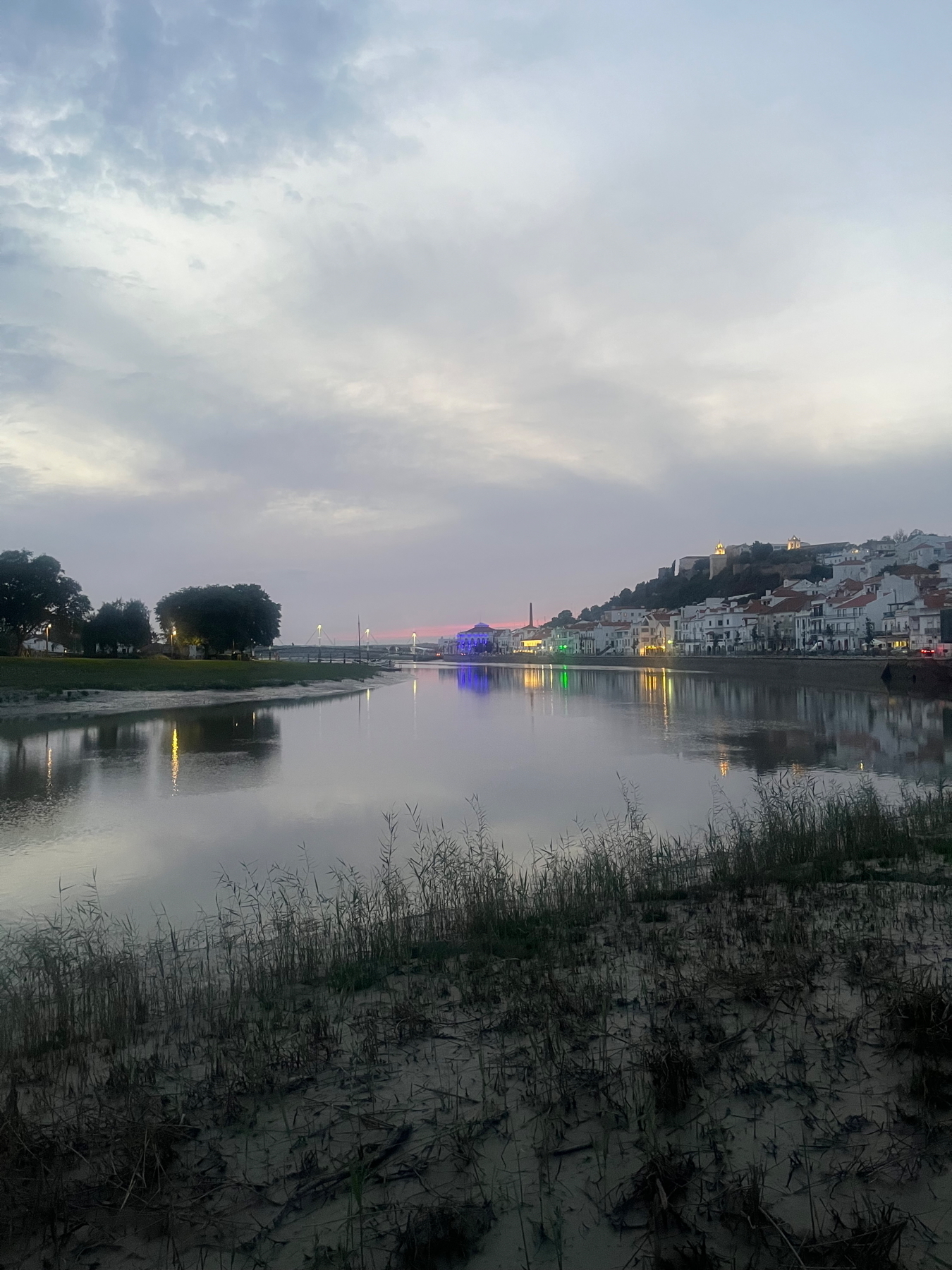 A serene riverside scene at dusk, with city buildings reflecting on the calm water. The sky is cloudy, and there are lights illuminating the area around the riverbanks and houses.