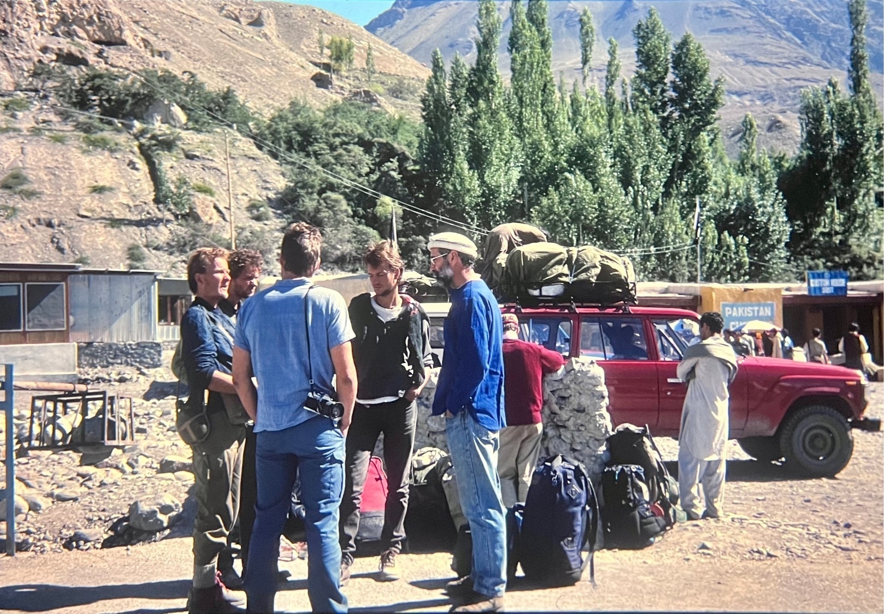 A group of western backpackers standing talking at the Pakistan border post, high in the dry mountains, a few trees and a red vehicle stand near by.