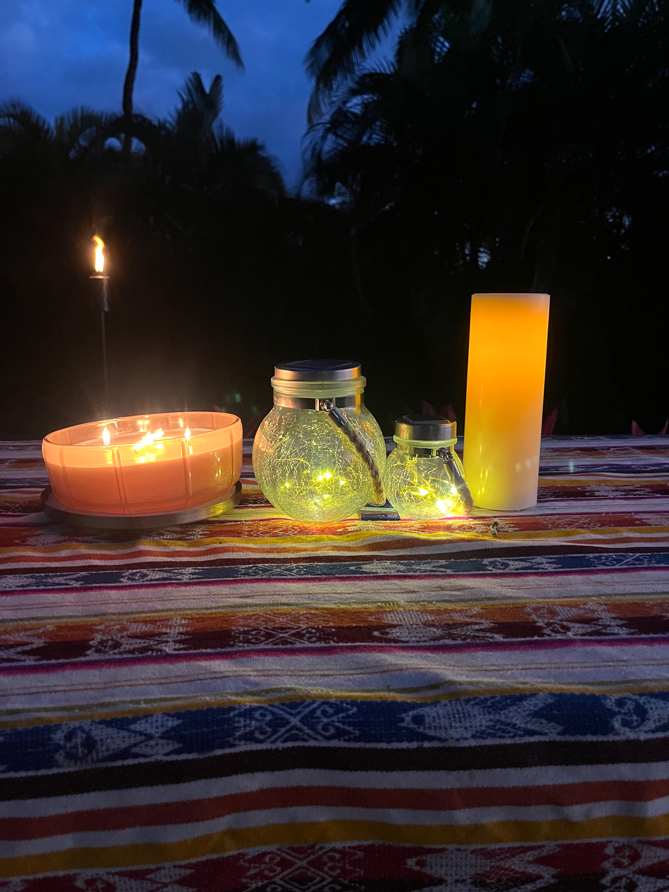 A collection of lit candles and string lights set on a vibrant, multicolored textile surface outdoors at dusk, with silhouettes of palm trees in the background.