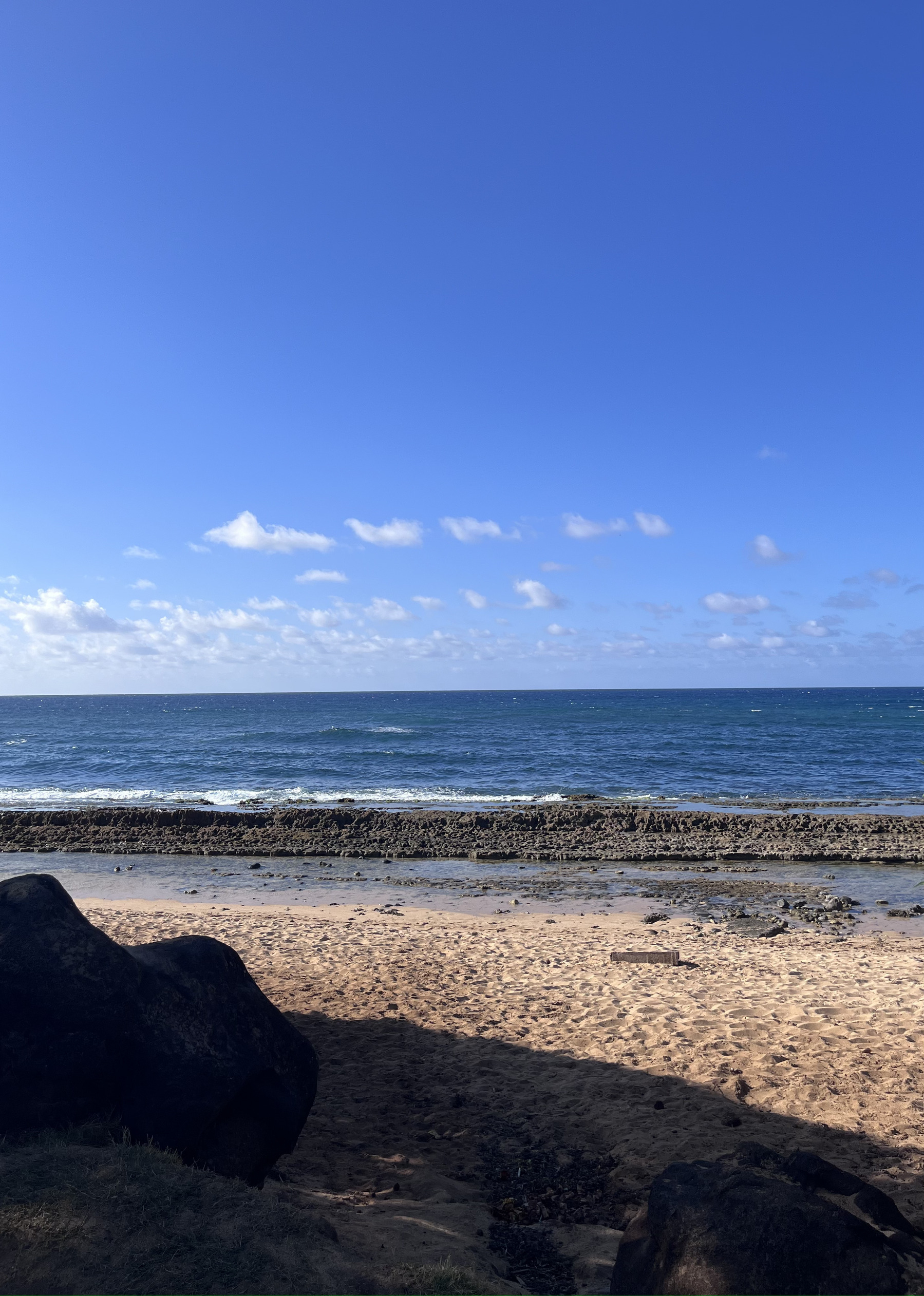 Auto-generated description: A serene beach scene shows a sandy shore, rocks, gentle waves, and a clear blue sky with scattered clouds.