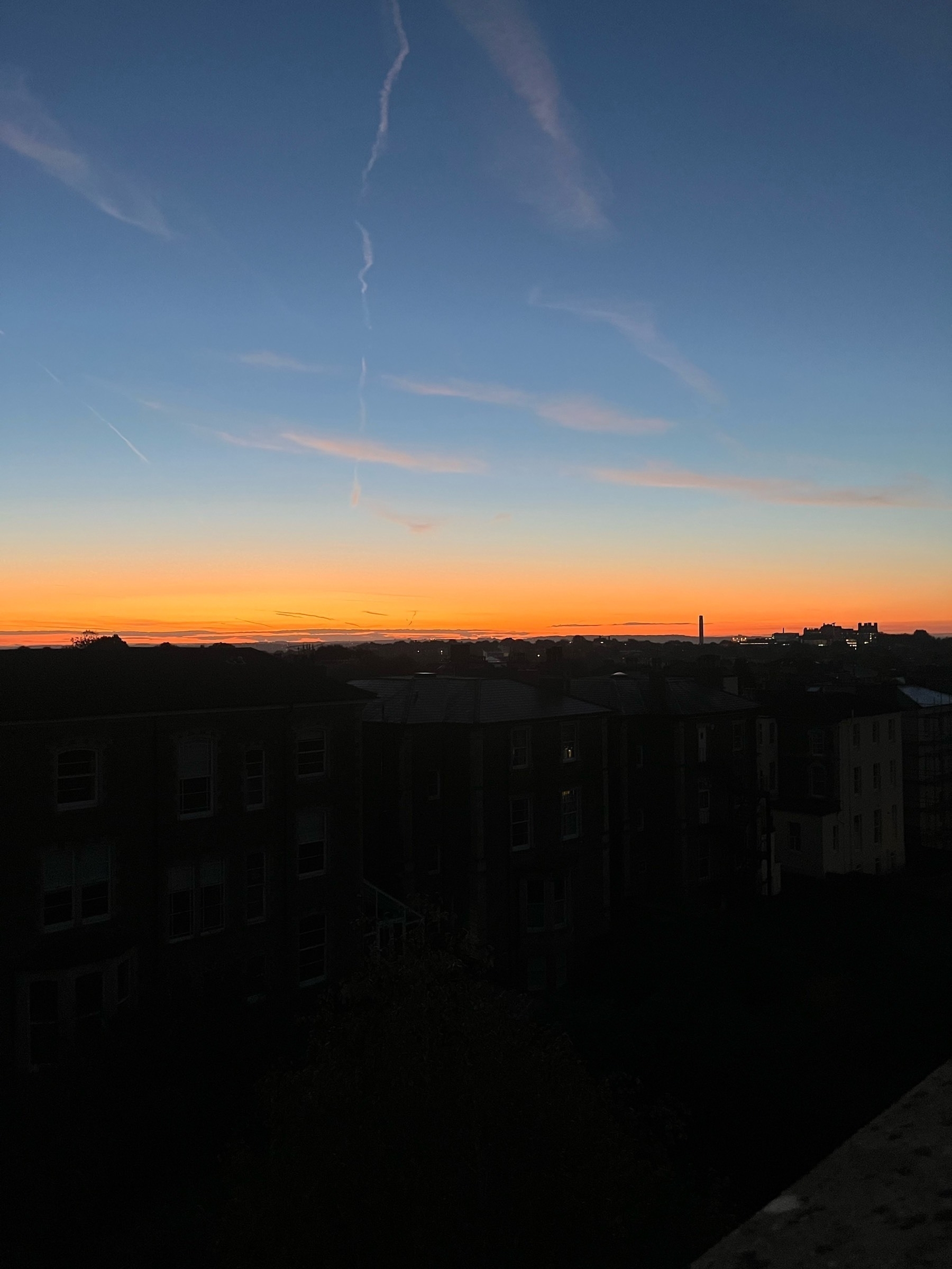 Red, orange and golden light on the horizon of a dark city (of Bristol) at dawn. Above the sky is blue with the trail of passing aircraft.