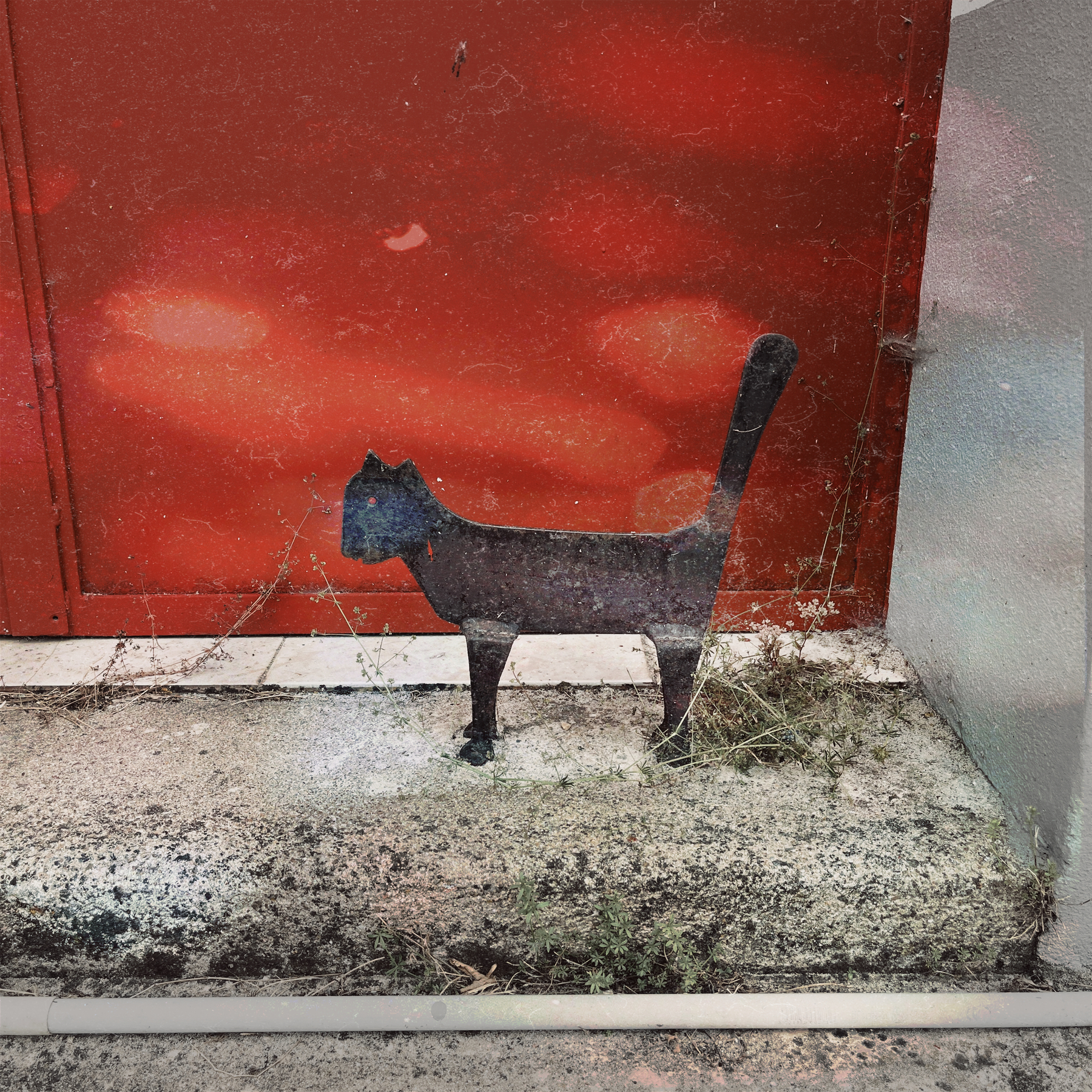 A flat metal statue of a black cat standing on concrete against a red door