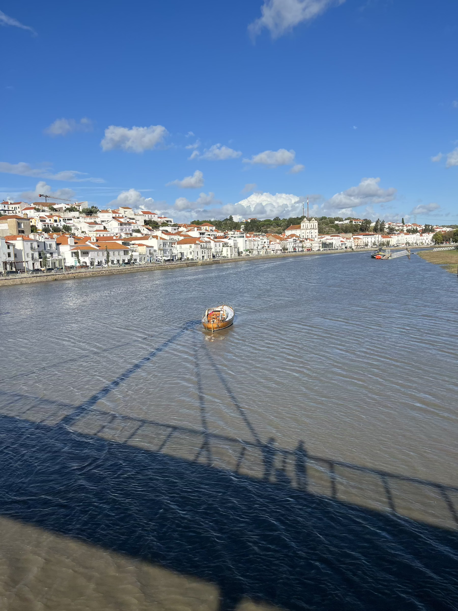 View from a bridge of a river with a boat moored to one side. A town of white houses rises up from the river. Above is a blue sky with scattered cloud. A reflection of the bridge with two people standing on it appears in the river.