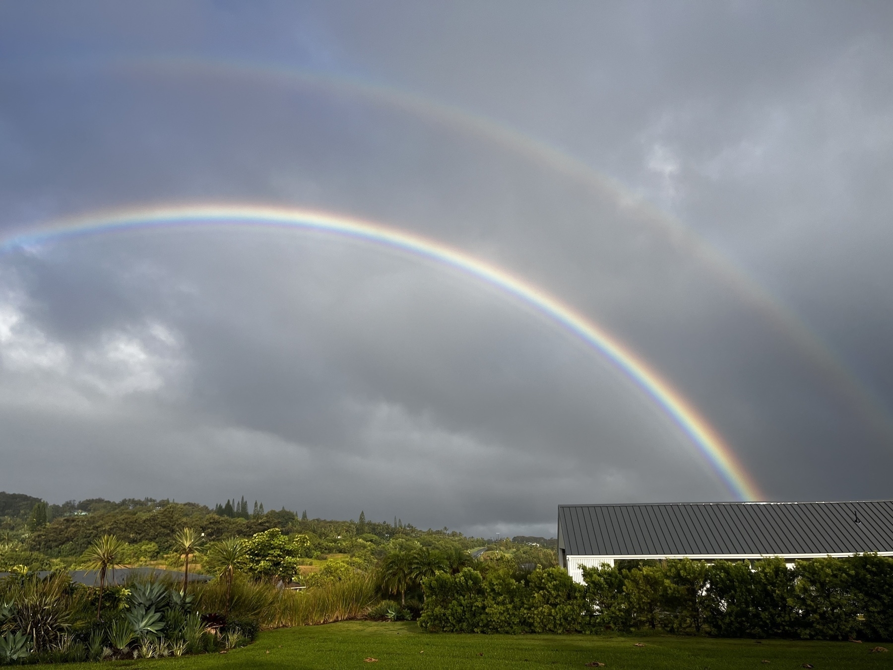 The arc of a double rainbow, one clearer than the other, appears out of house roof over green vegetation, against an overcast sky.