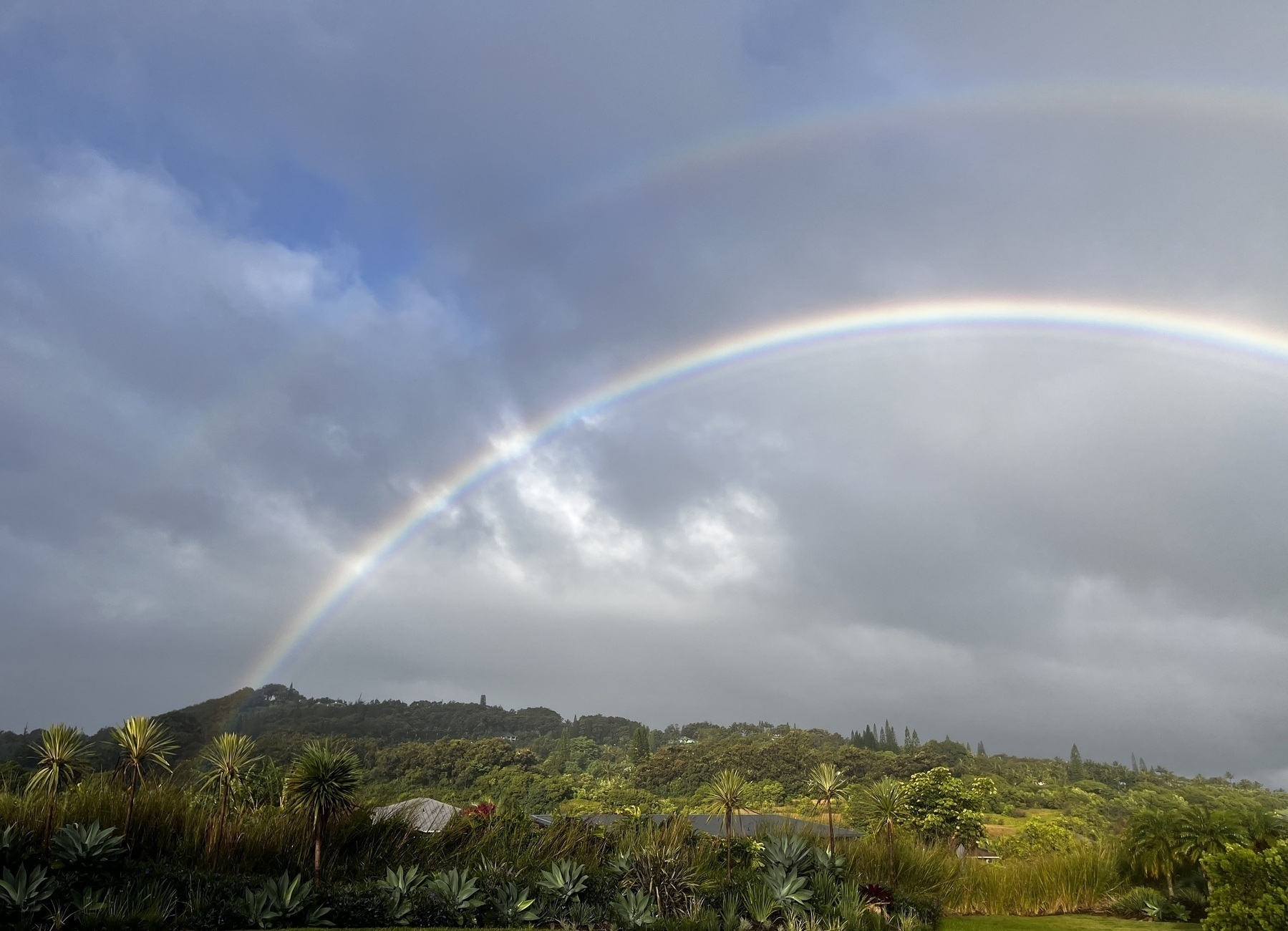 An arc of a double rainbow, one clearer than the other, appears over green hillsides and concealed rooftops, again an overcast sky with patches of blue sky.