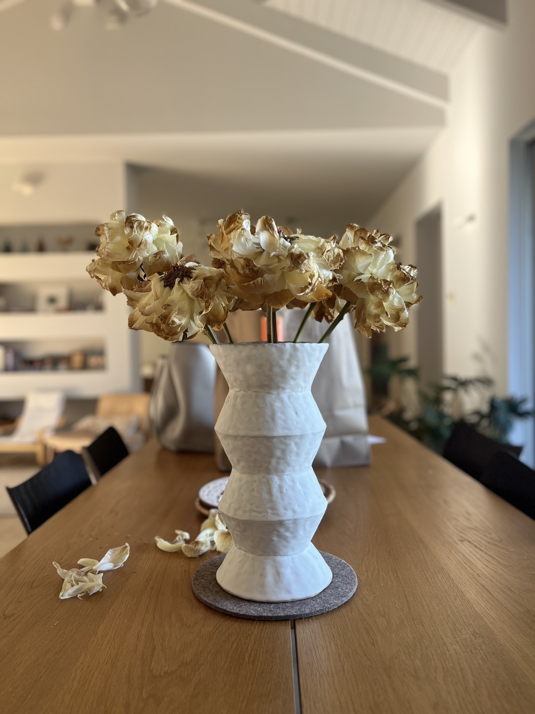 A white vase with dying white and brown peony flowers sits on a wooden table in a modern, minimalist room