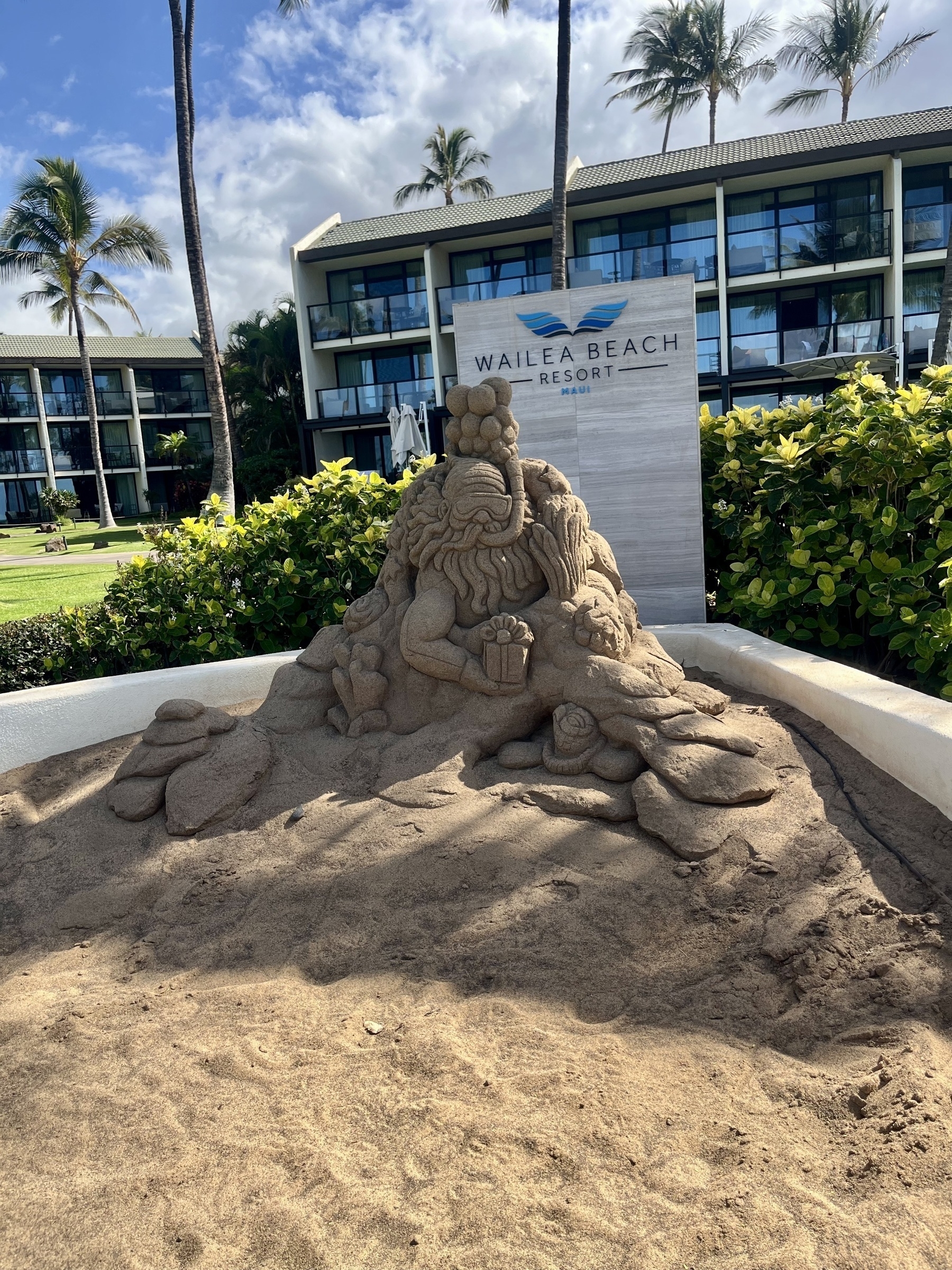 A detailed sand sculpture of a sea creature wearing a crown is displayed near a resort sign with palm trees and a hotel building in the background.