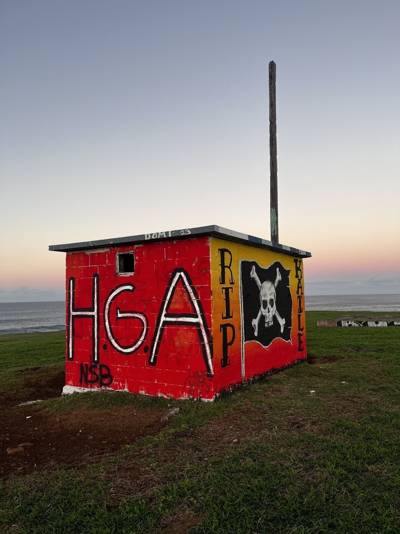 Two sides of a small brick building in a field near the ocean is covered in graffiti. The side nearest the camera is red with large white letters spelling HGA. The other side, against a gradient background from yellow at the top to read at the bottom, has a large skull and crossbones flag with the text RIP on one side and KAILE.