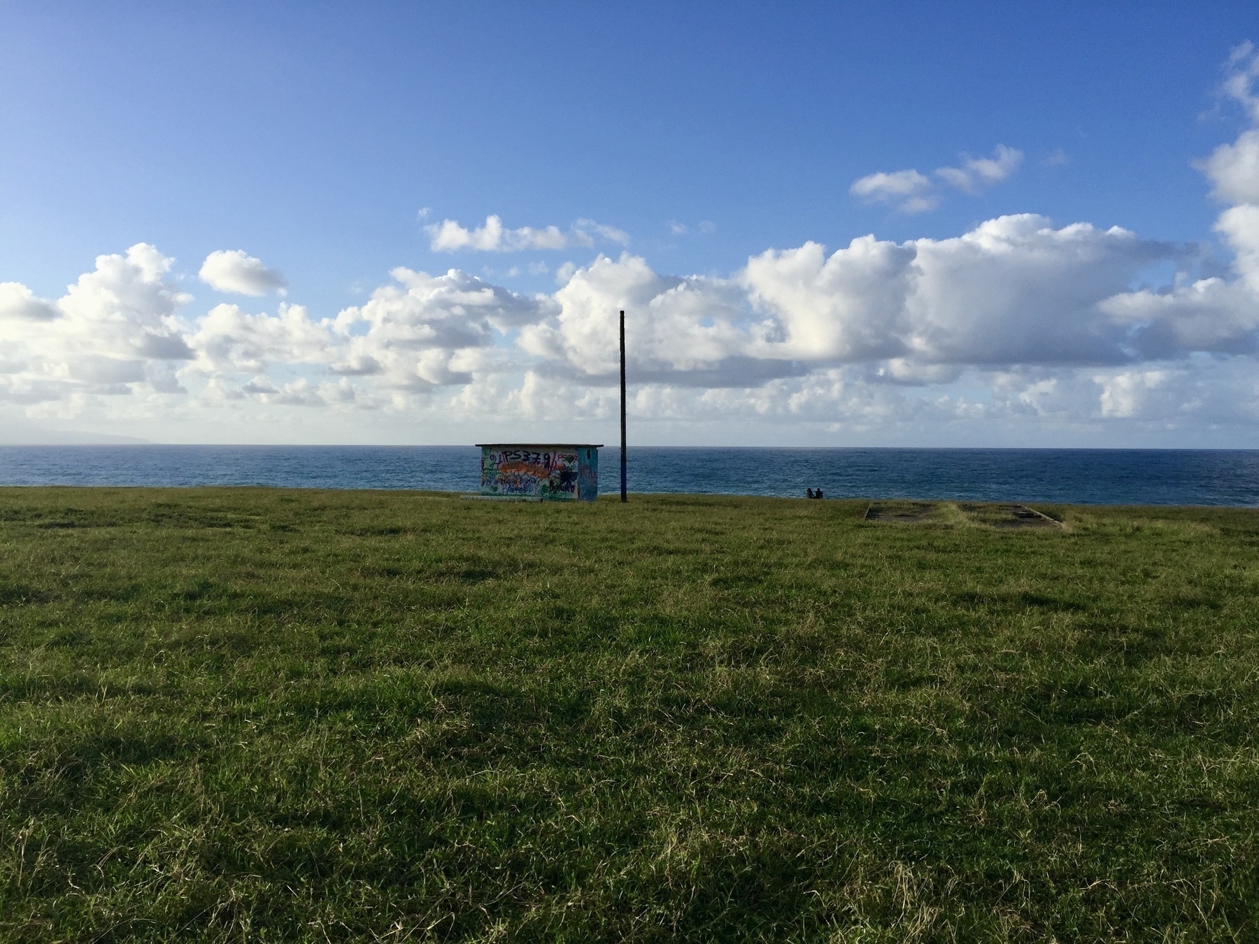 A grassy field stretches to the ocean under a blue sky with scattered clouds, with a small, graffiti-covered structure sitting near the edge of the field near to the ocean with a wooden pole next to it. Two people sit on the edge go the field looking out to the ocean. The overgrown foundations of another building can be made out in the field to the left of the building.