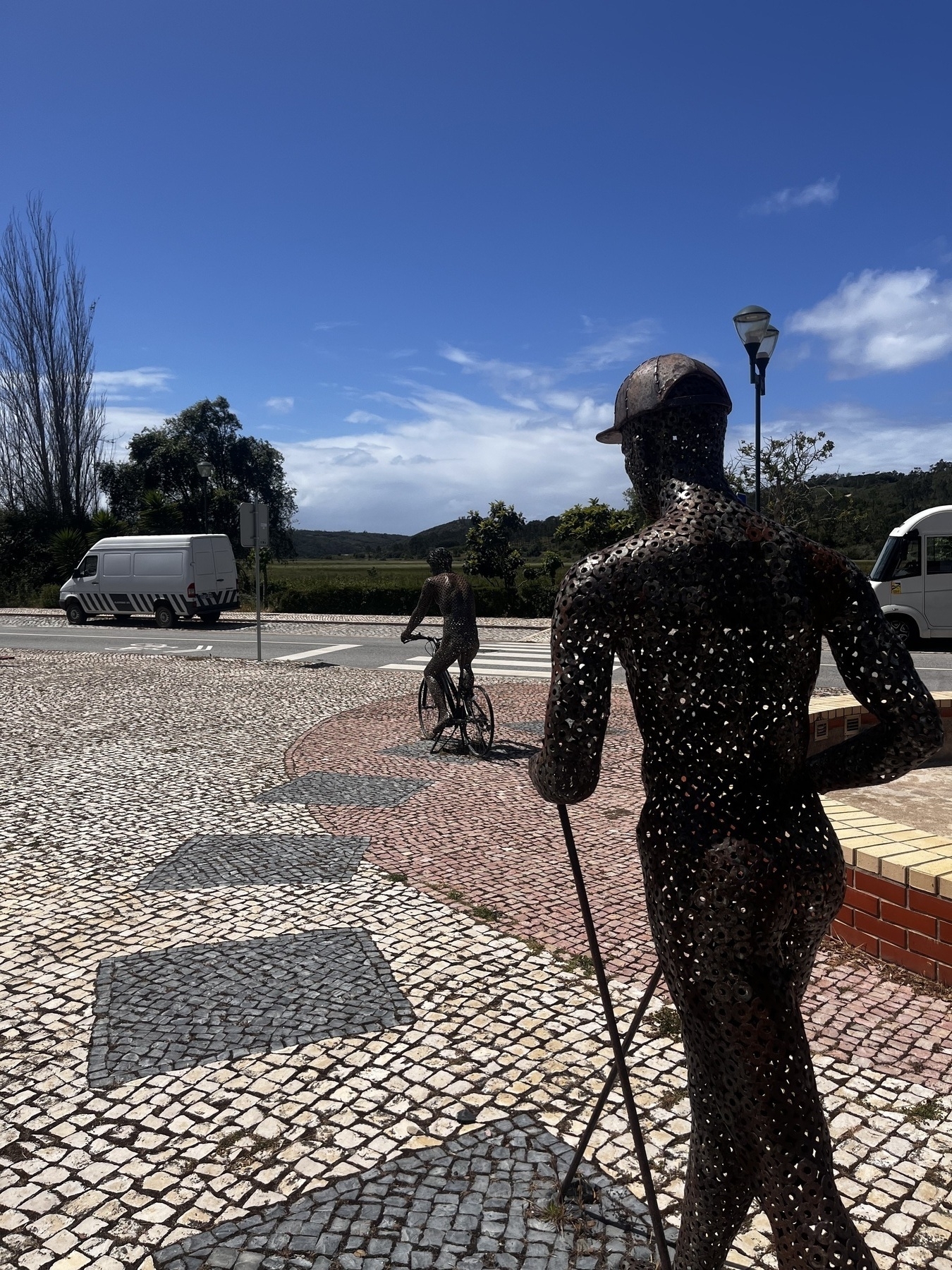 Metal statues of a person walking and cycling in a small square on the edge of a town.