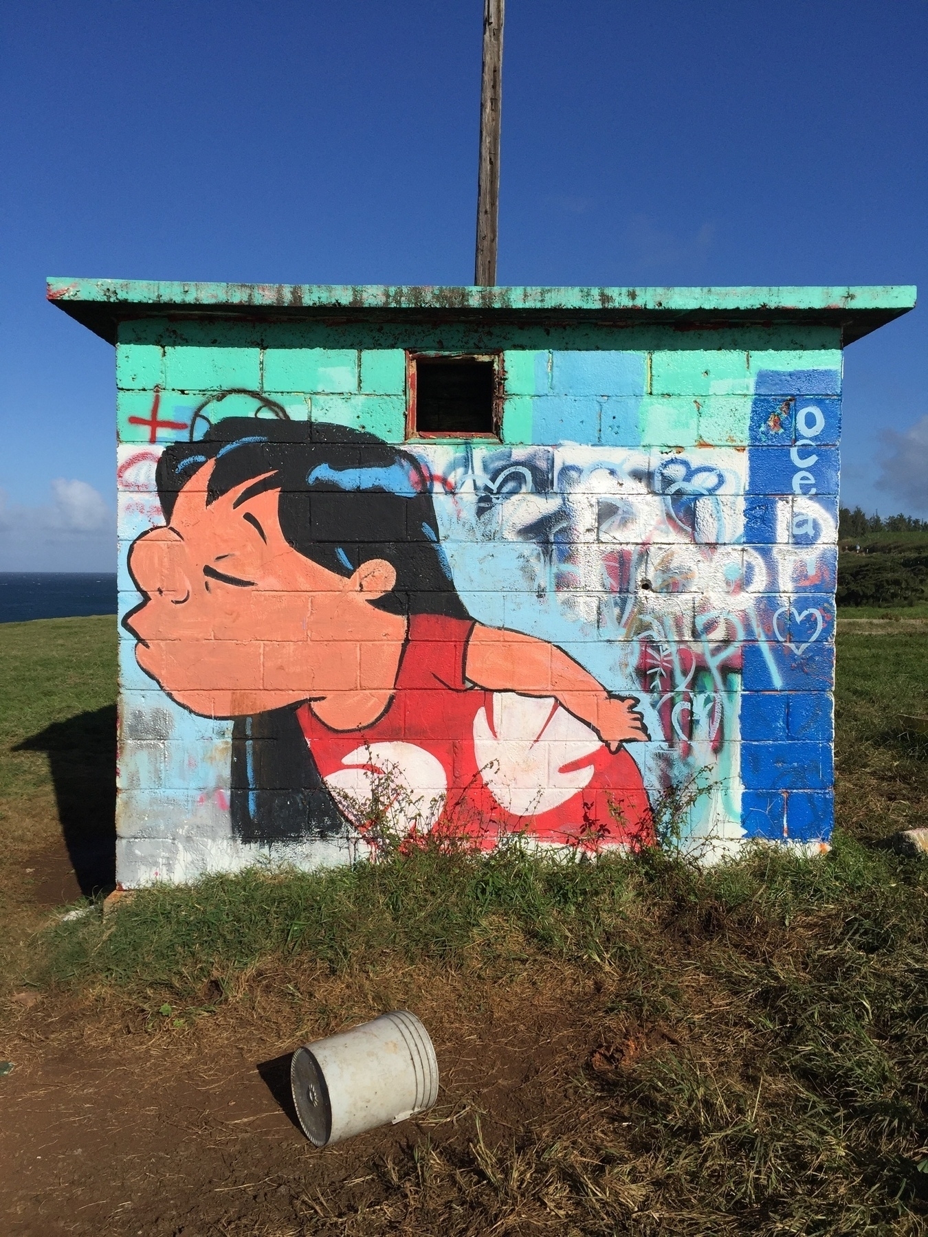 A graffiti of a cartoon character in a red shirt is painted on a small, blue, brick structure with a bucket on the ground nearby. The ocean is visible in the left background.