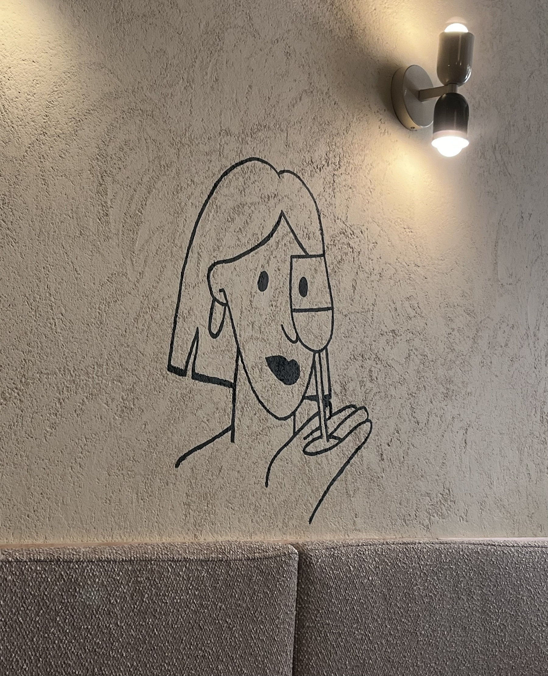 Drawing on a wall in a restaurant of a woman eying a glass of wine. A double light shines above