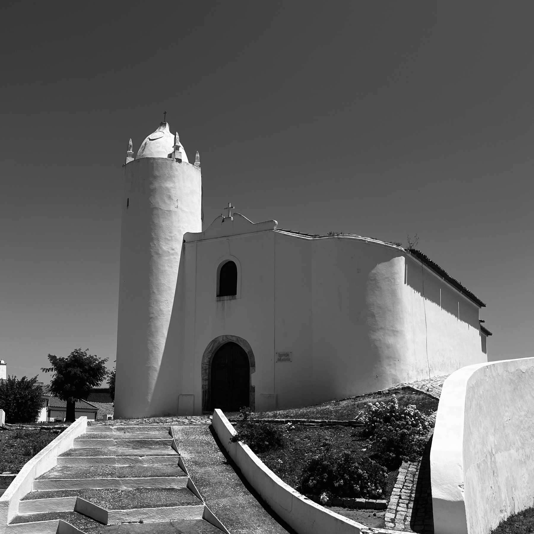 A black and white photo of a simple white church, a round tower on one side, steps leading up to it and a curved wall below and on the right