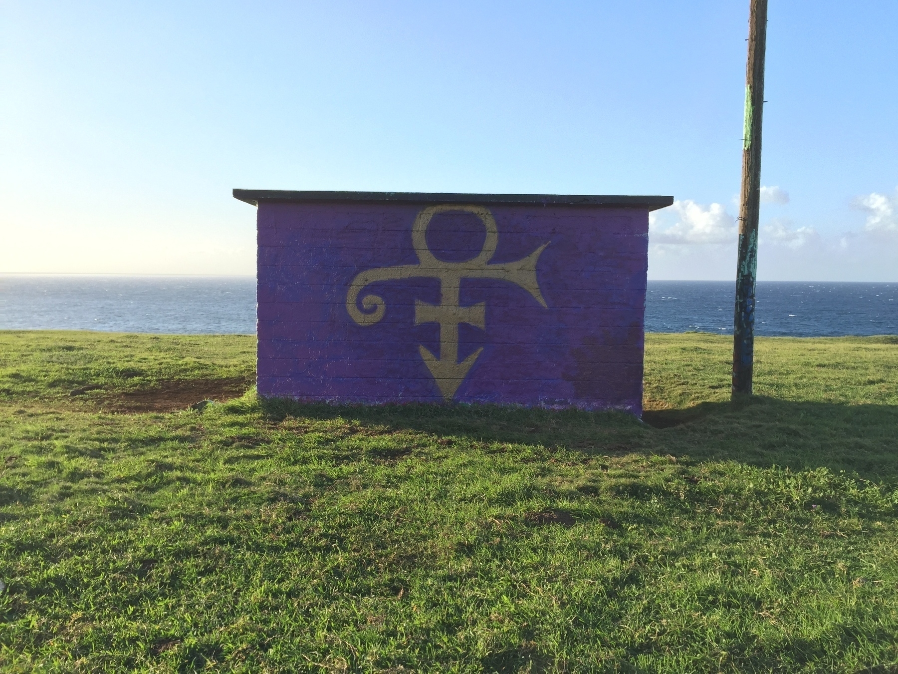 A brick building standing in a field with the ocean behind it and a wooden pole to its right. The bulding is painted with a purple background and a symbol that was associated with the pop star Prince.