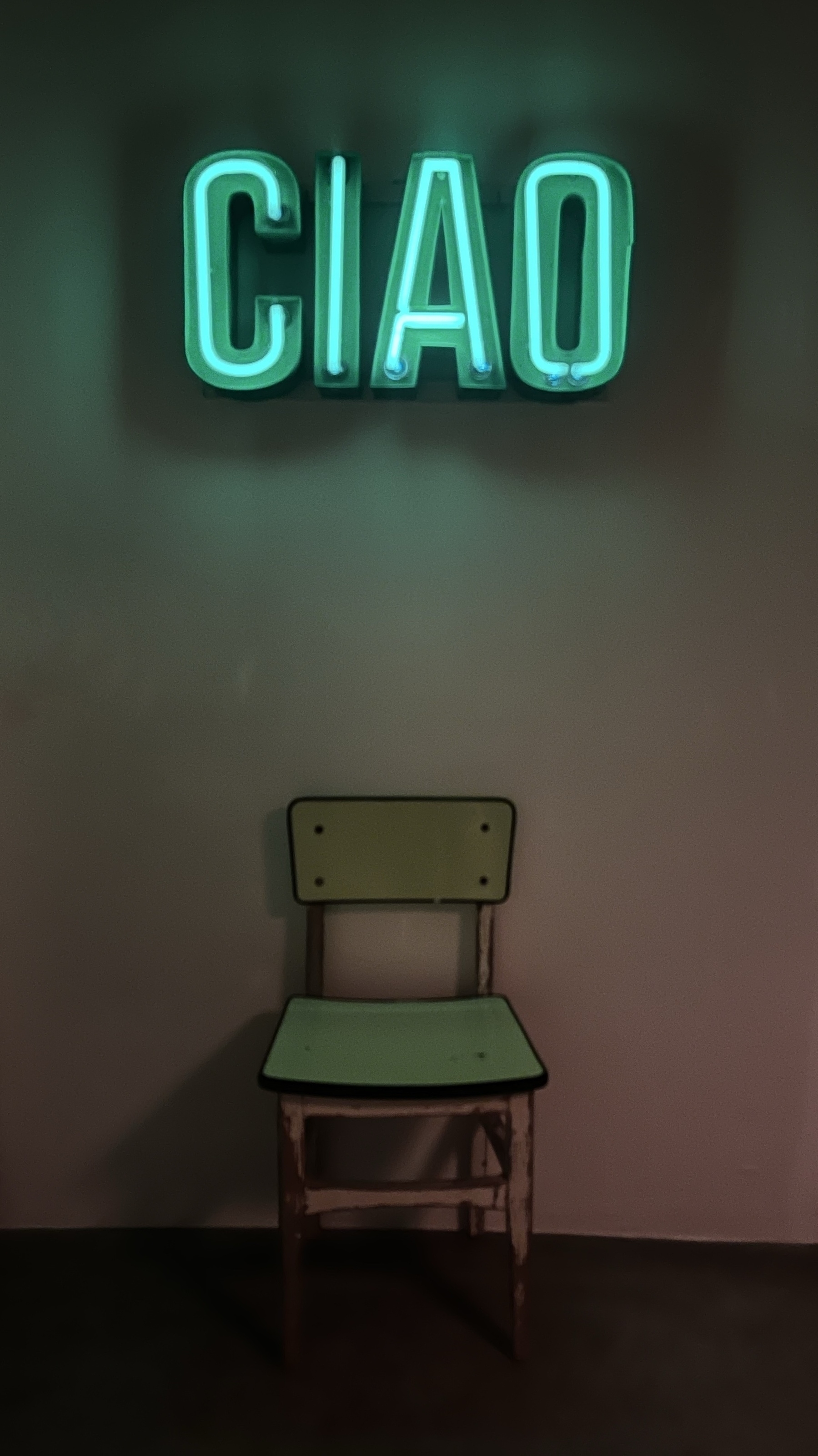 A green neon sign spelling out CIAO hanging on a white wall. Below is a simple chair with a green back and green seat.