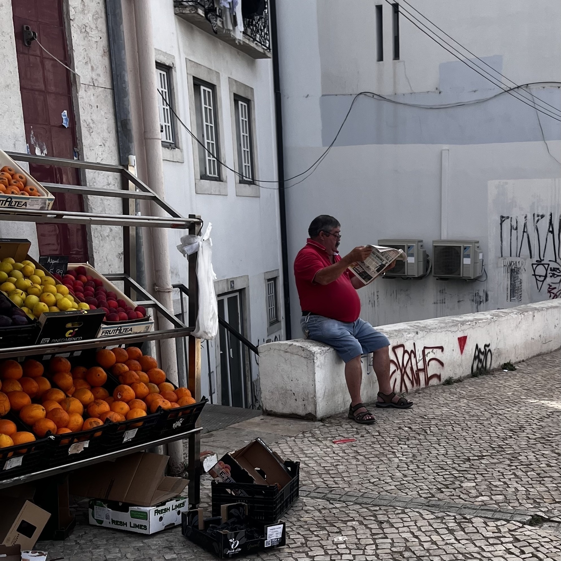 A man sits on a small wall reading his newspaper. He is wearing a red t-shirt which covers a bulging belly. He wears glasses,has a mustache, blue denim shorts and sandals. Beside him is a fruit stand.