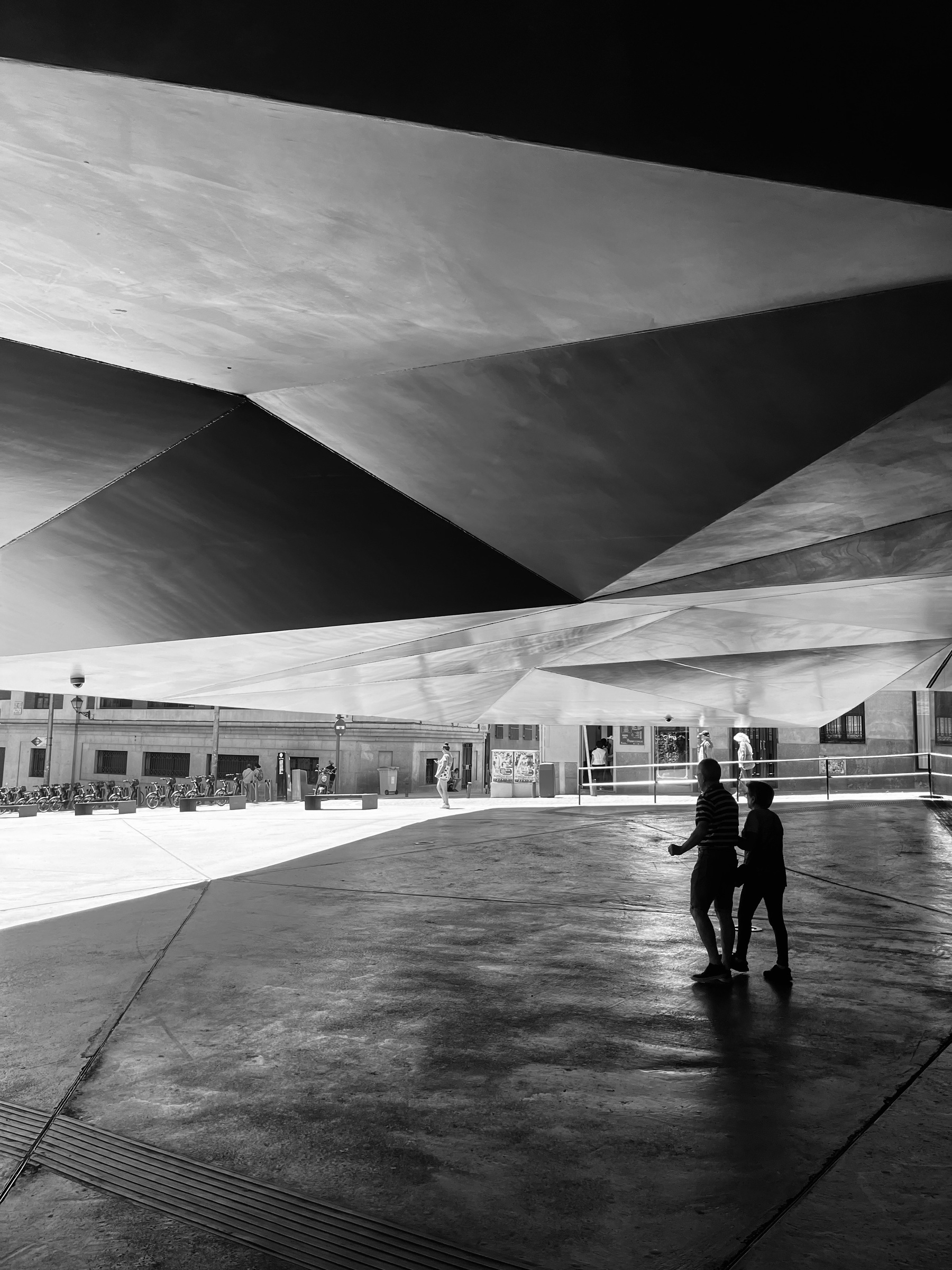 A black and white photo showing a couple walking under a low roof made up of shapes, in the sun away from the building people walk around.