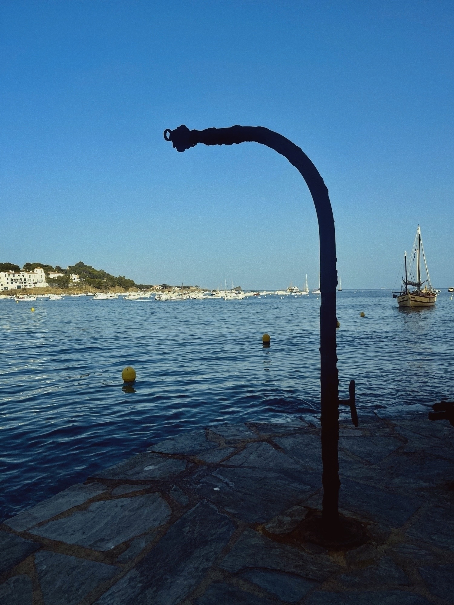 A metallic “L” shaped object standing upright on the water’s edge. Behind is an ocean bay with an old sail boat anchored off shore
