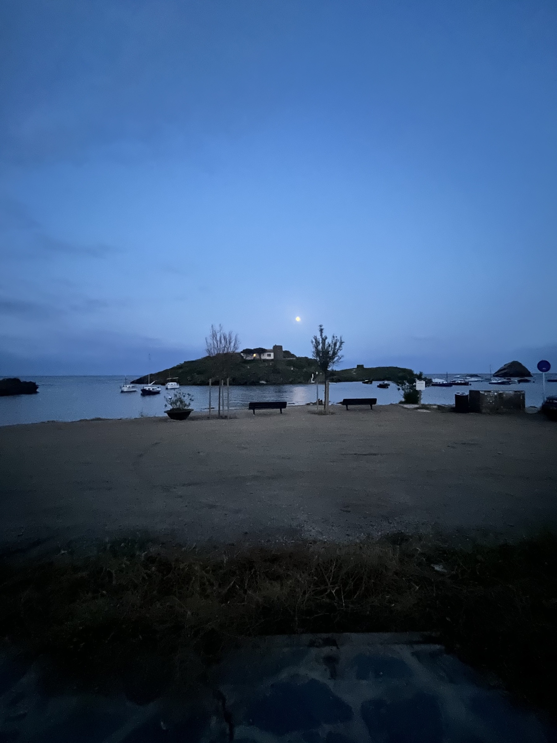 A neat full moon rising over a small island with a building on it that include a turret. Small craft boast dot in the calm water. In the foreground is a smooth dirt road with two trees and two chairs at the water’s edge.