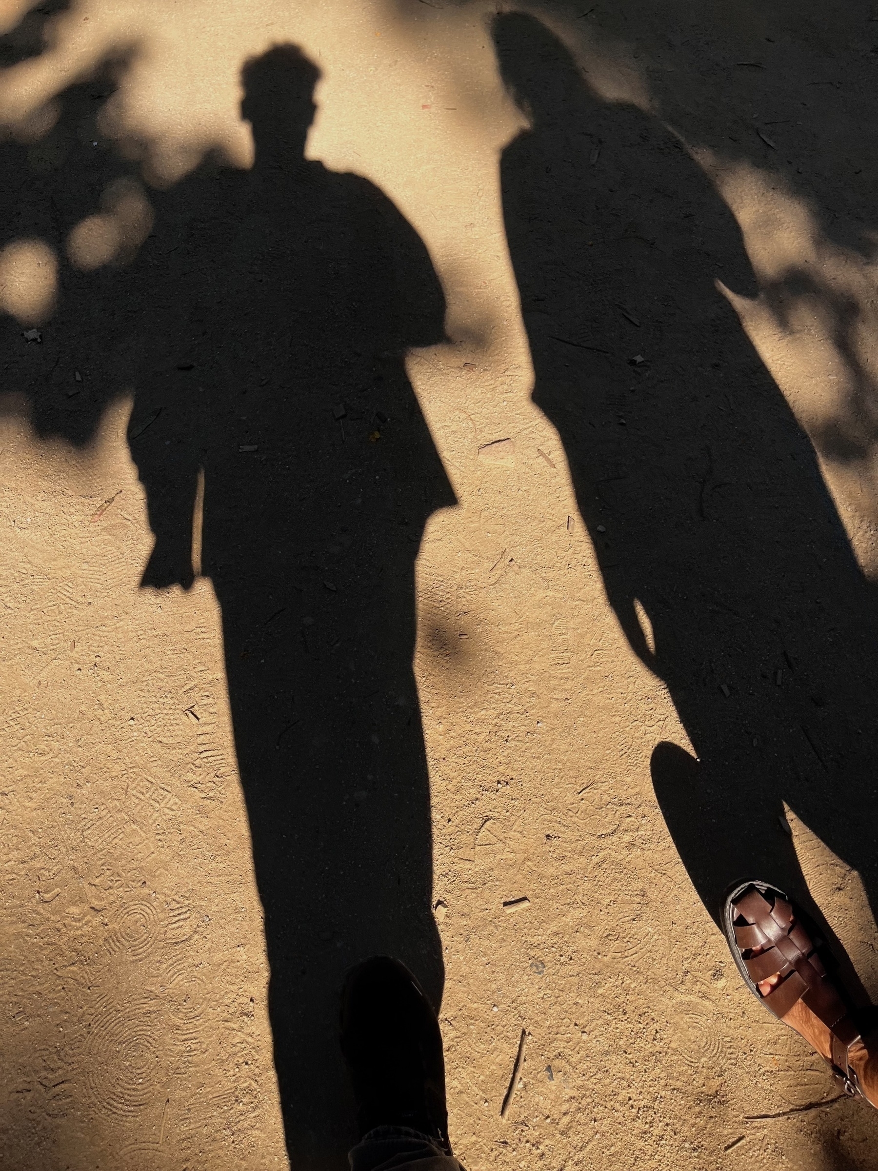 The shadow of two people walking on hard sandy coloured surface. A foot of each person is visible.