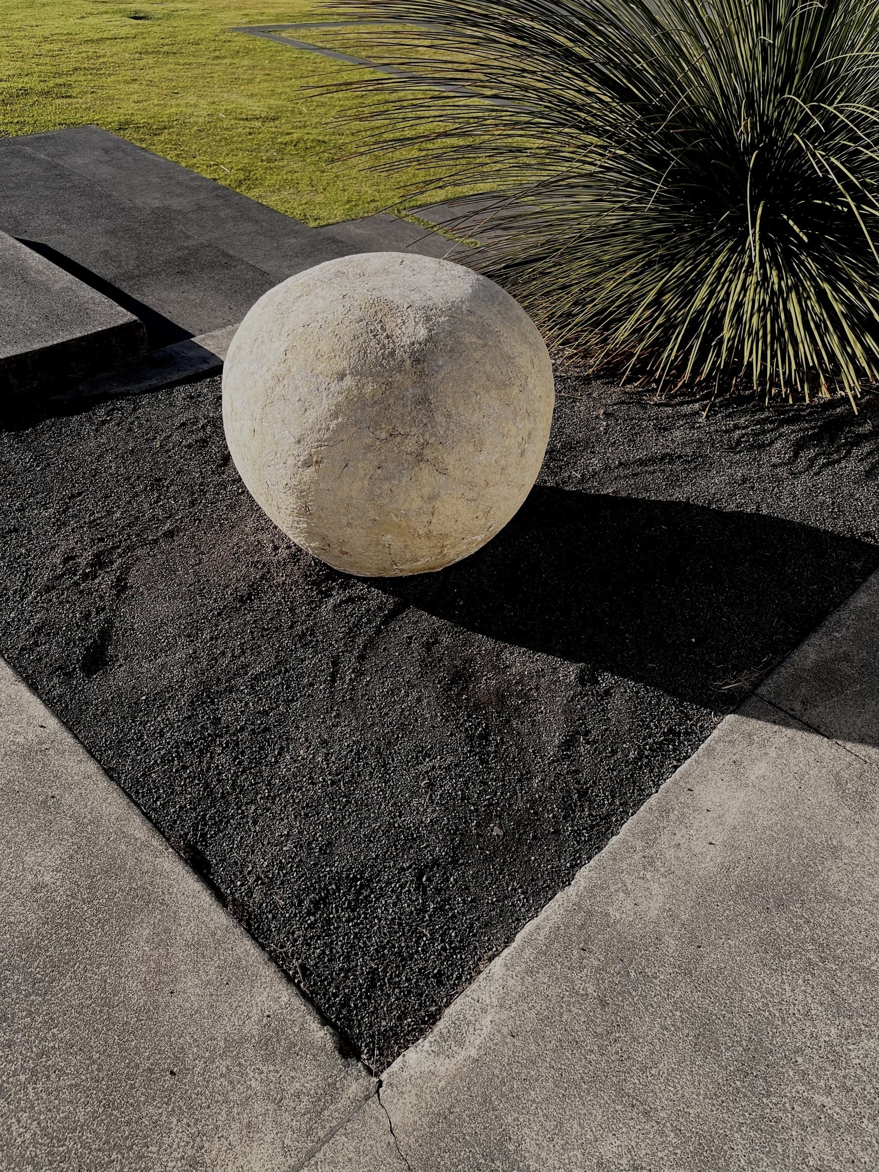 A large stone sphere sits on a landscaped area with dark gravel, concrete pathways, and greenery in the background.