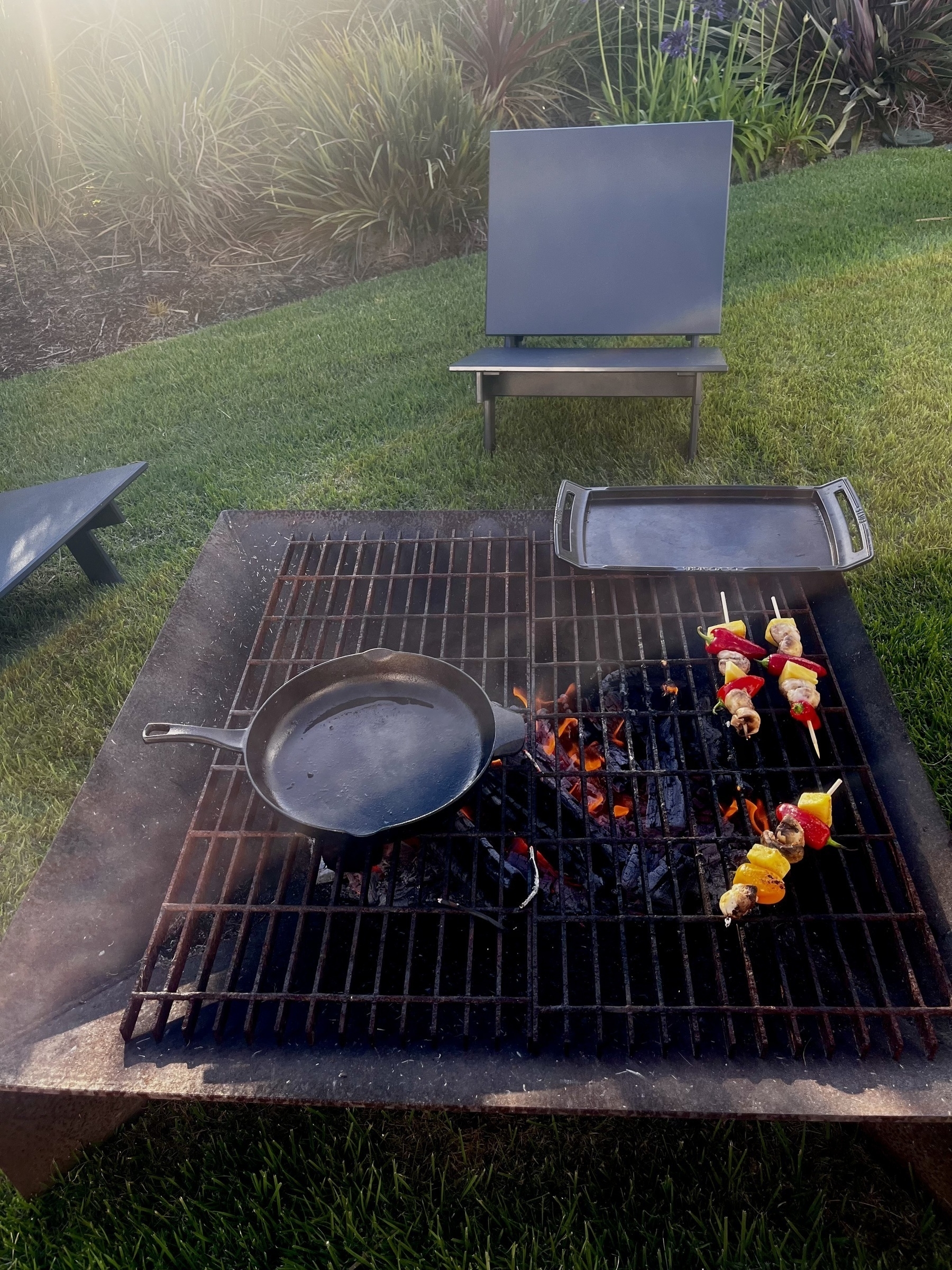 A grill with a pan and skewers of meat and vegetables is set up outdoors on a grassy area with chairs in the background.