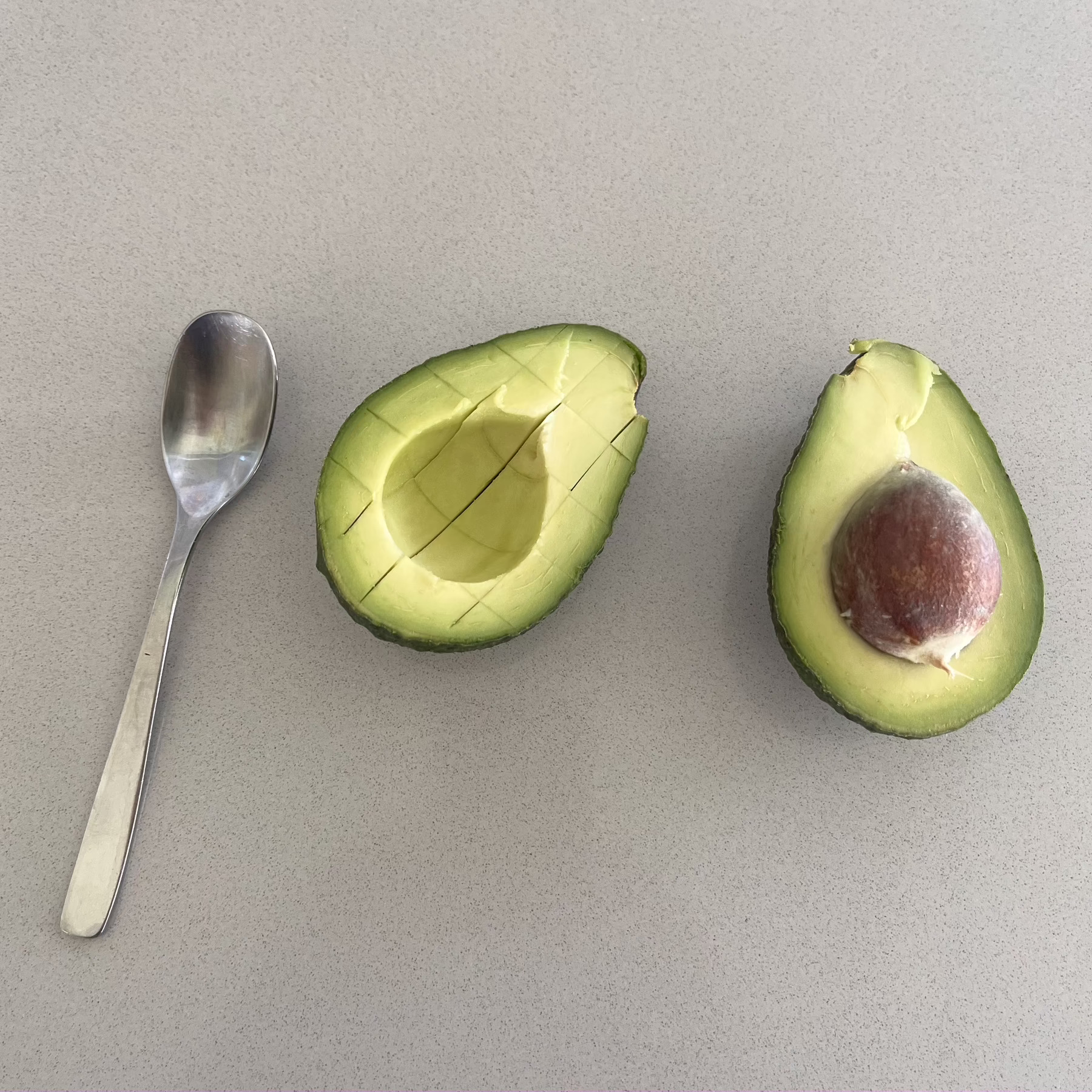 An avocado split in two and lying facing up on a counter top. One half has a pip in it. A small silver spoon lies beside the fruit.