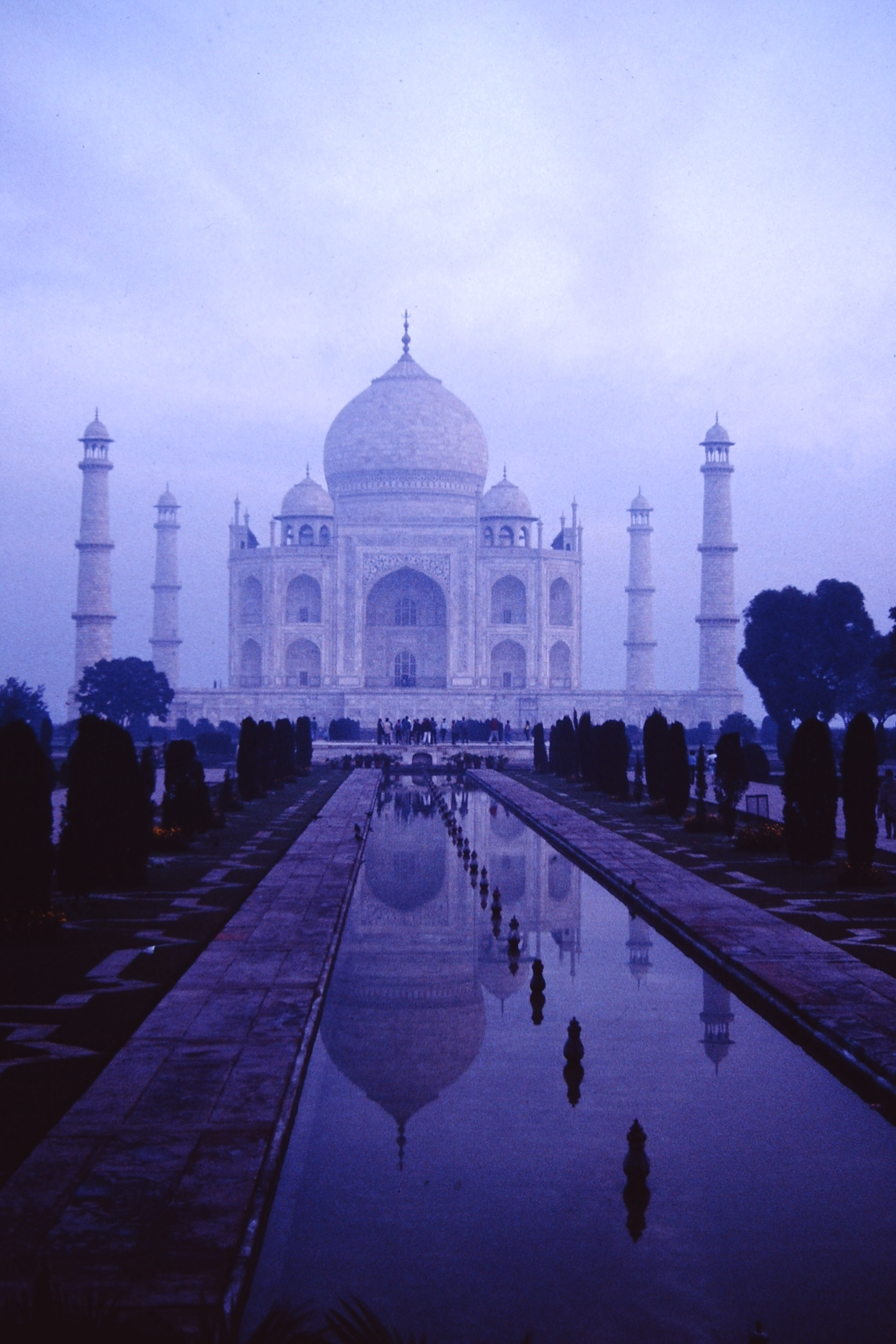 The Taj Mahal, an ivory-white marble mausoleum built in the tradition of traditions of Indo-Islamic and Mughal architecture seen on a misty morning across its reflecting pool.