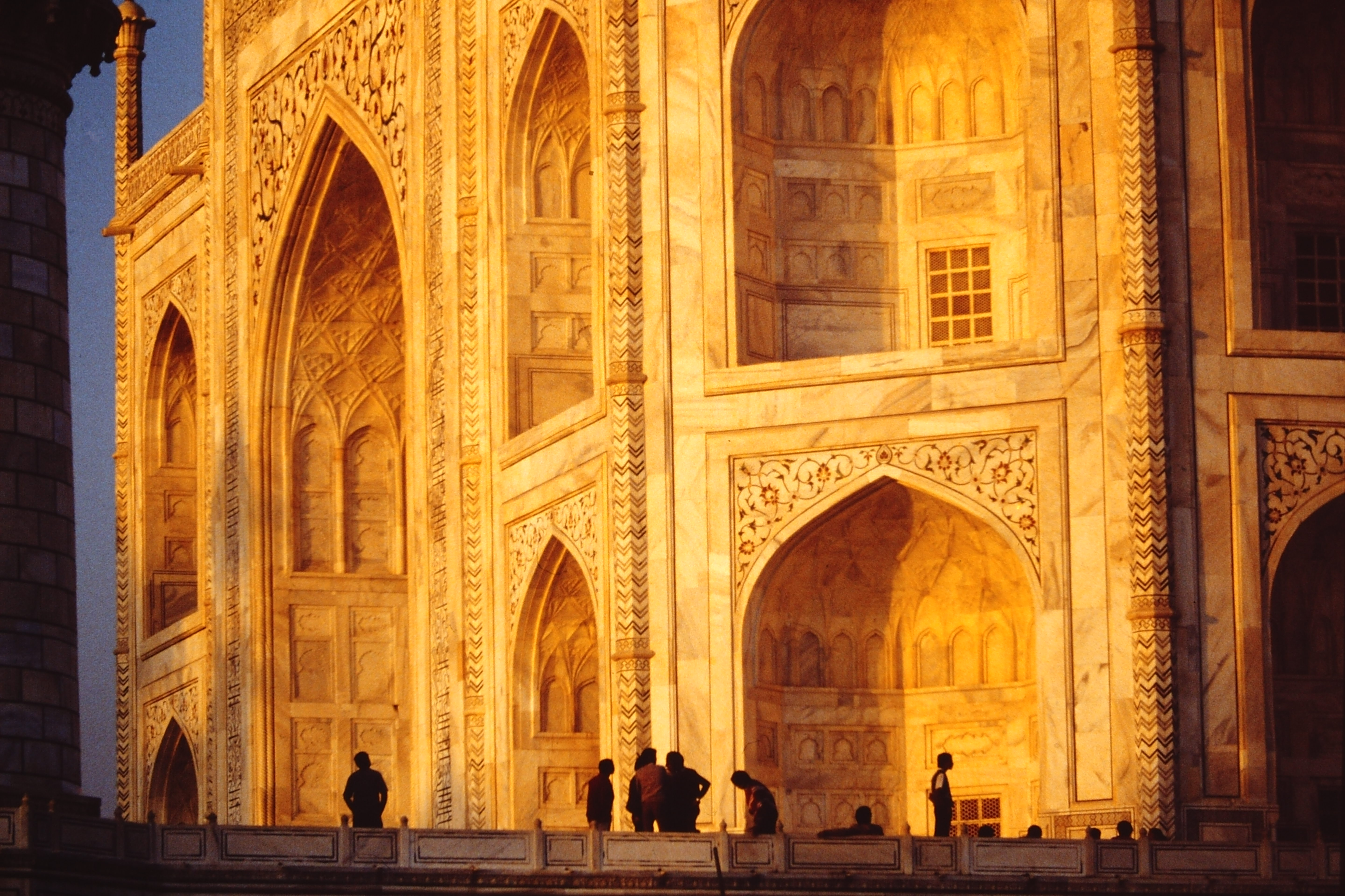 A close up of the marble side of the Taj Mahal with the sun shining on it creating a gold glow, with people walking around in front.