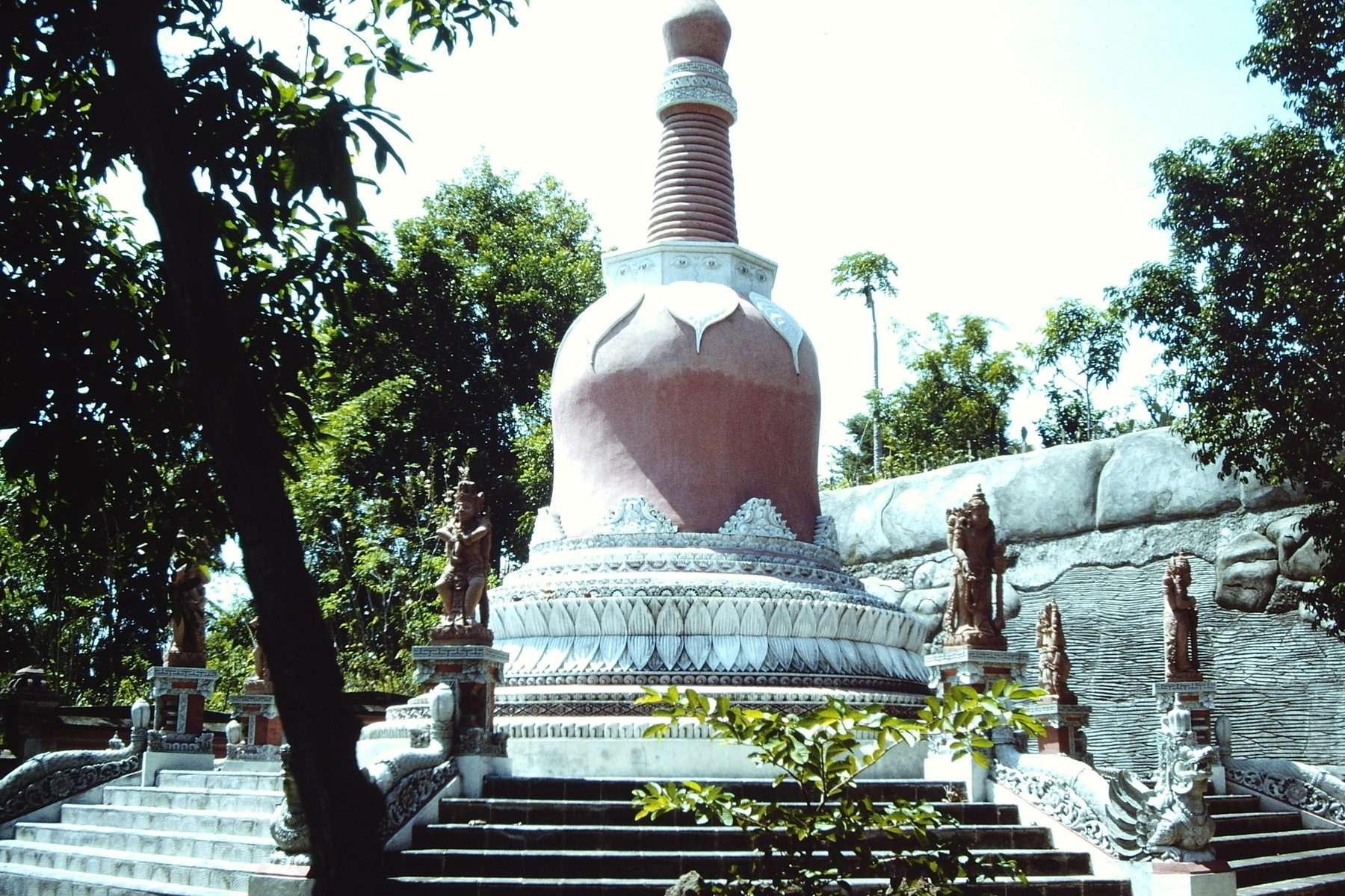 A large stupa surrounded by Balinese statues and trees is depicted, set against a backdrop of greenery.