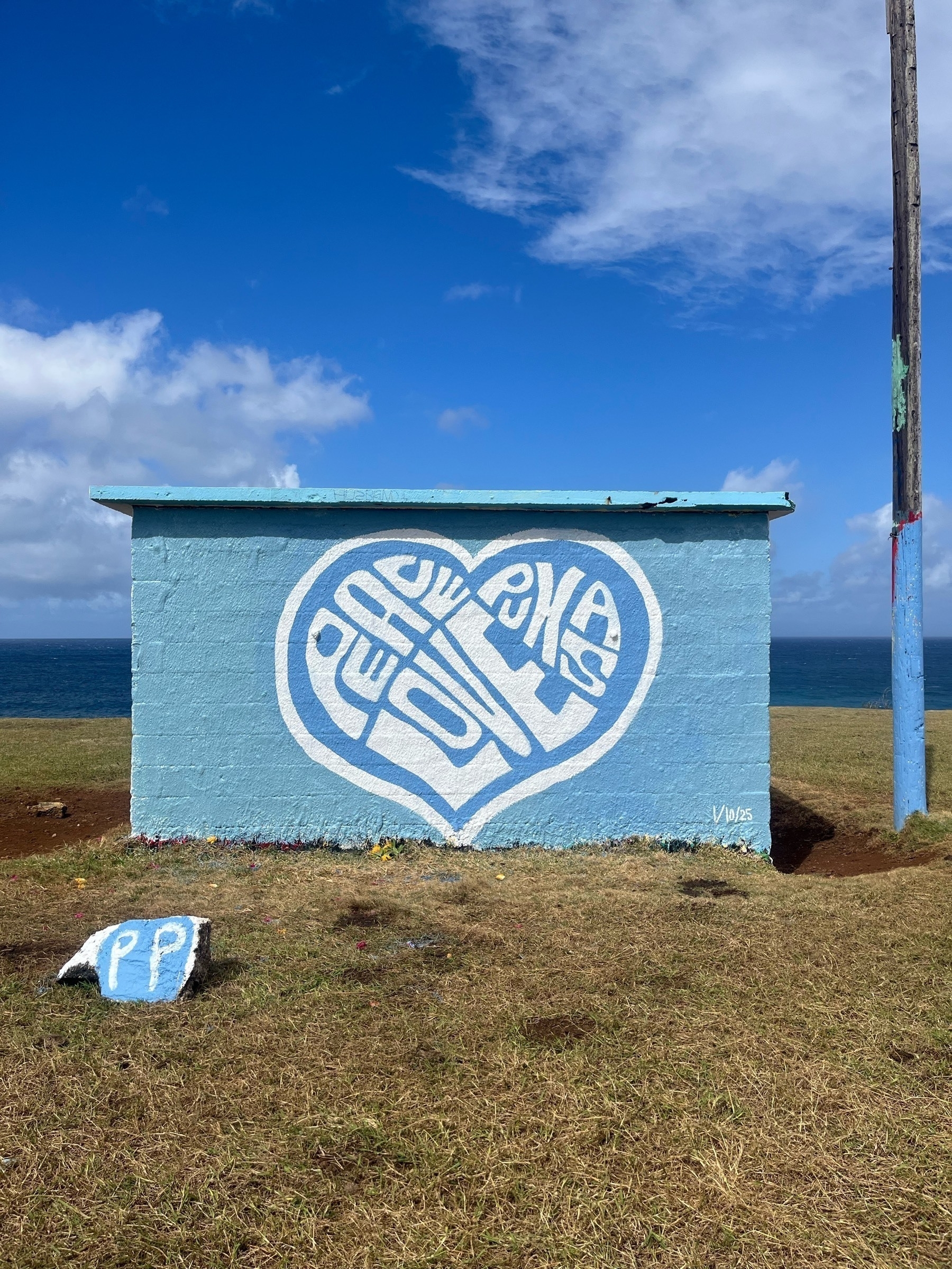 A small blue building by the ocean features a large white heart with the words Peace Love Pukas