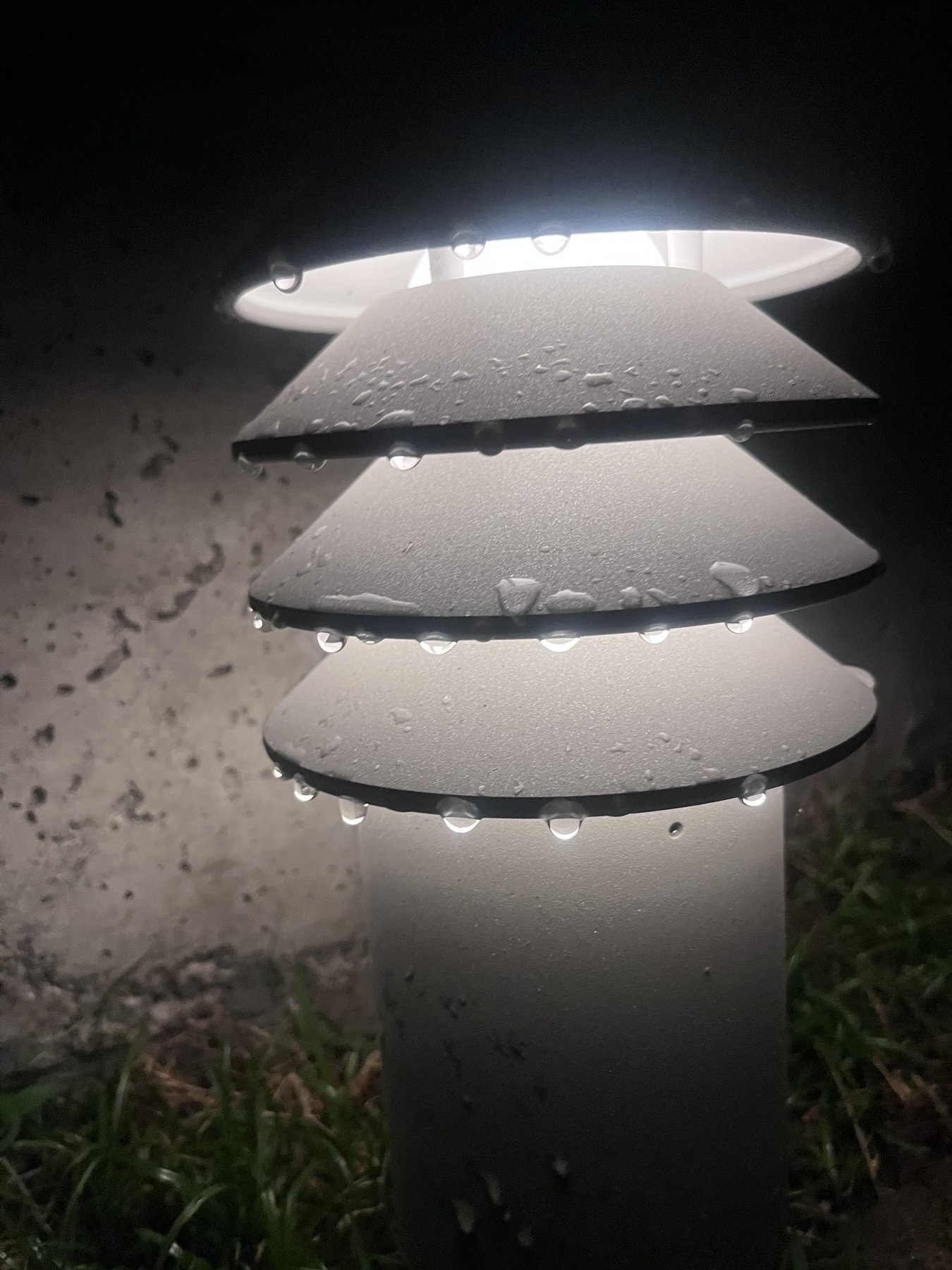 An outside light with multiple layers. Rain droplets hang from each layer. The light is on. Visible behind the light is concrete, and below grass. 