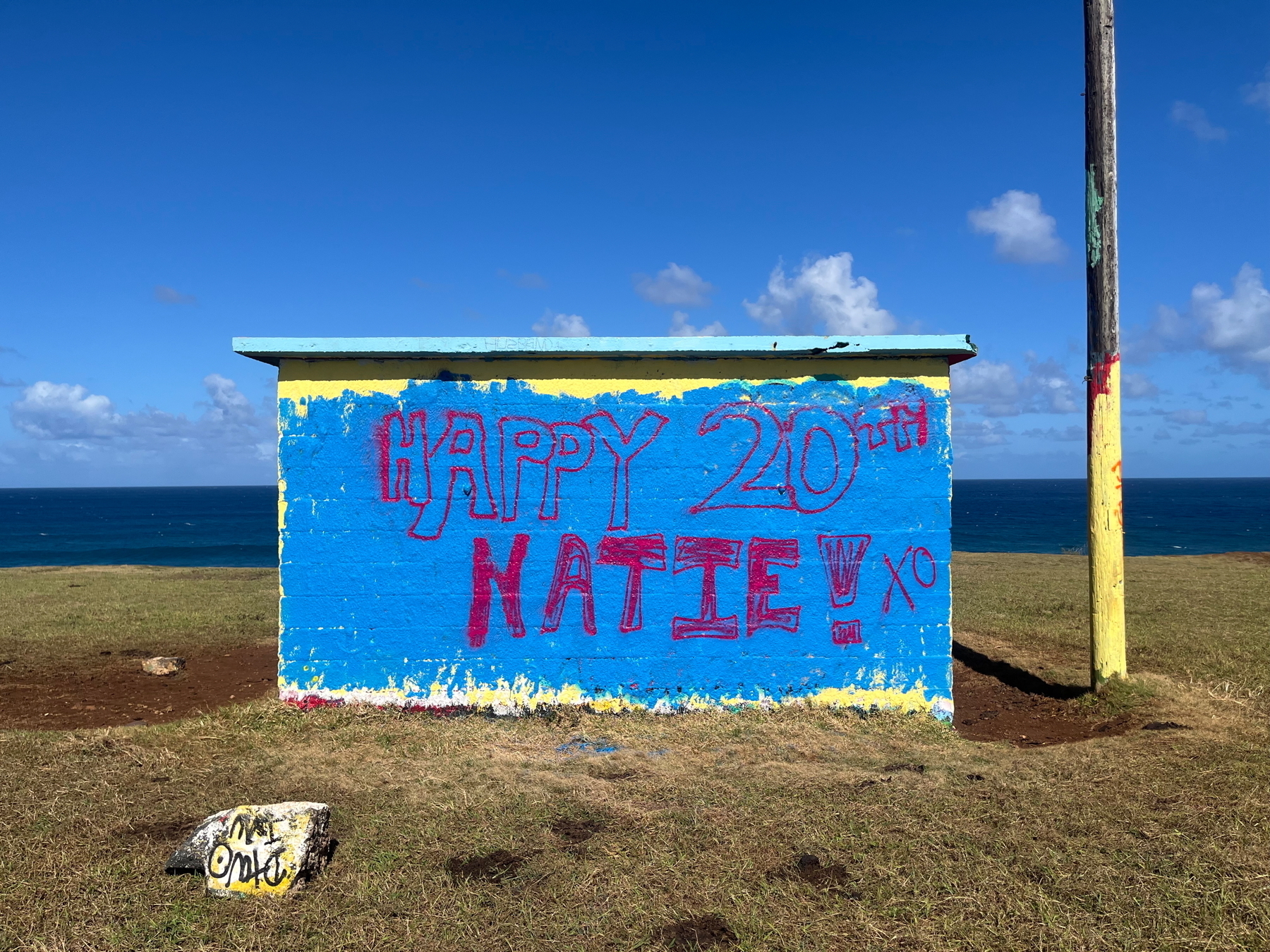 A small brick building painted blue, near the ocean displays a hand-painted message celebrating Happy 20th Katie! with a bright sky and grassy foreground.