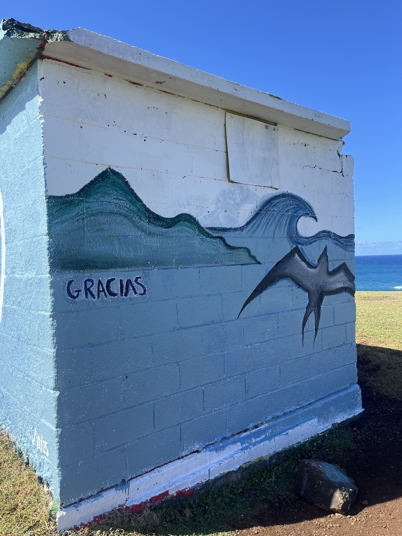 A small building features a mural of ocean waves, an island and a bird, and the word Gracias. The sky is white and the rest in shades of blue. A glimpse of the ocean can be seen on the right hand side.