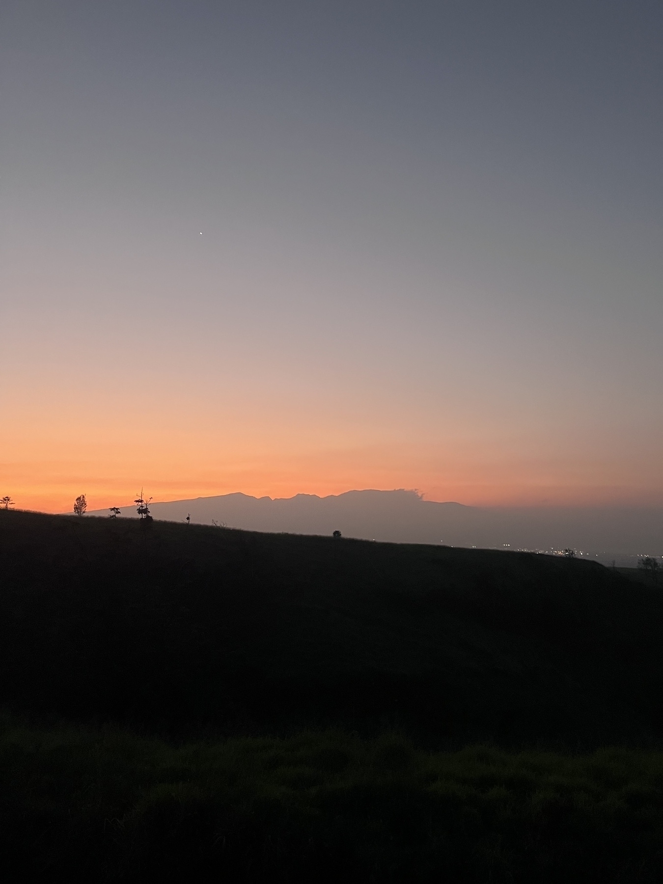 A peaceful landscape at dusk features a glowing orange horizon over a silhouette of the West Maui Mountains and a faintly visible cityscape.