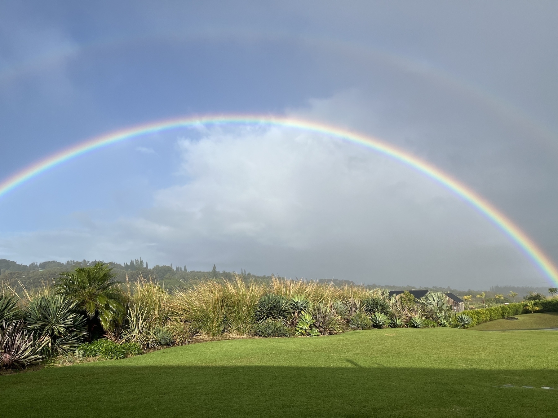 A vibrant rainbow arches over a lush green landscape on a partly cloudy day. A second faint rainbow can be seen above it.