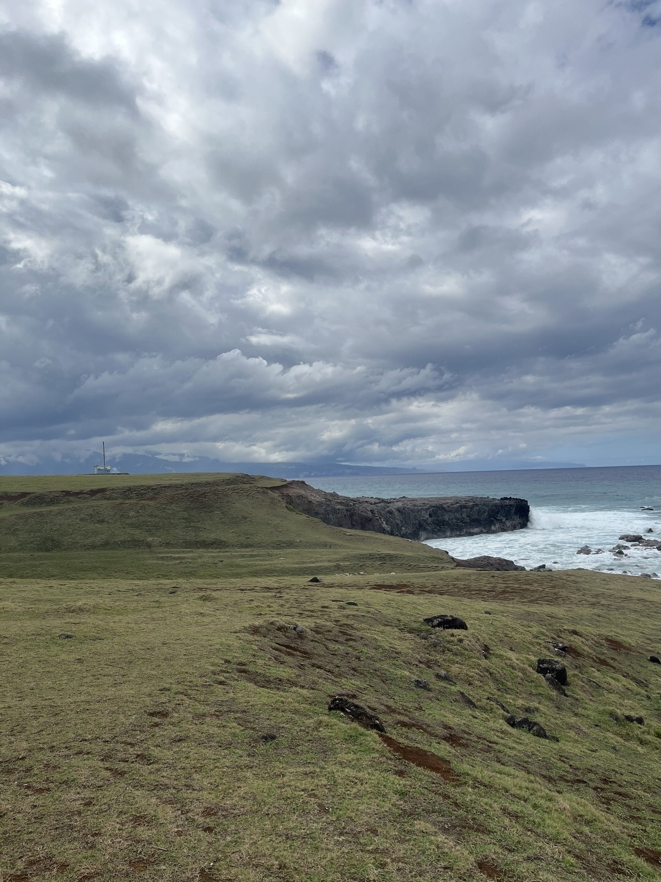 A grassy landscape meets rocky cliffs by the ocean under a cloudy sky. On the top of the cliffs sits a small brick building with a wooden post next to it. Another landmass disappearing into the clouds is visible in the distance. Another island is faintly visible in the further distance.