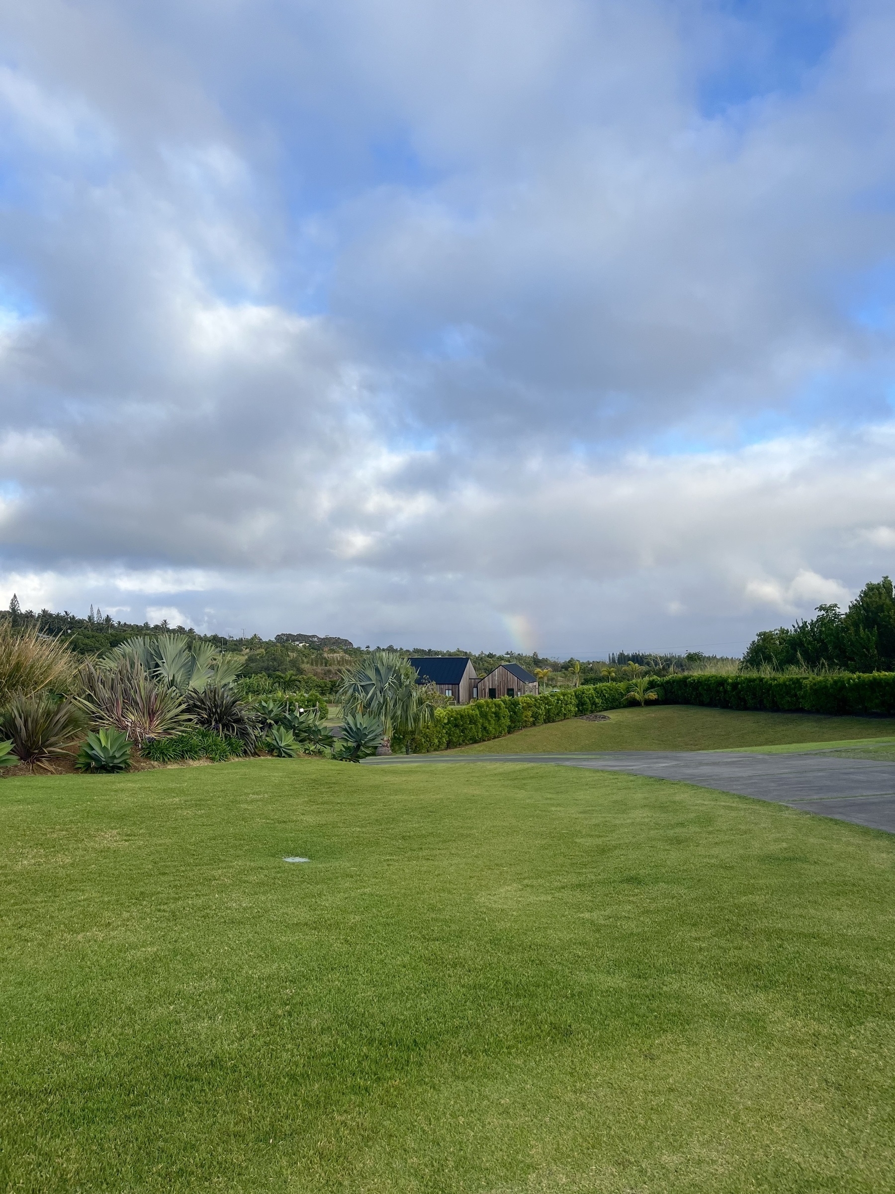 A lush, green lawn extends towards distant hills under a partially cloudy sky, with a small structure visible in the background. Behind that, out in the ocean, is a small, faint rainbow.