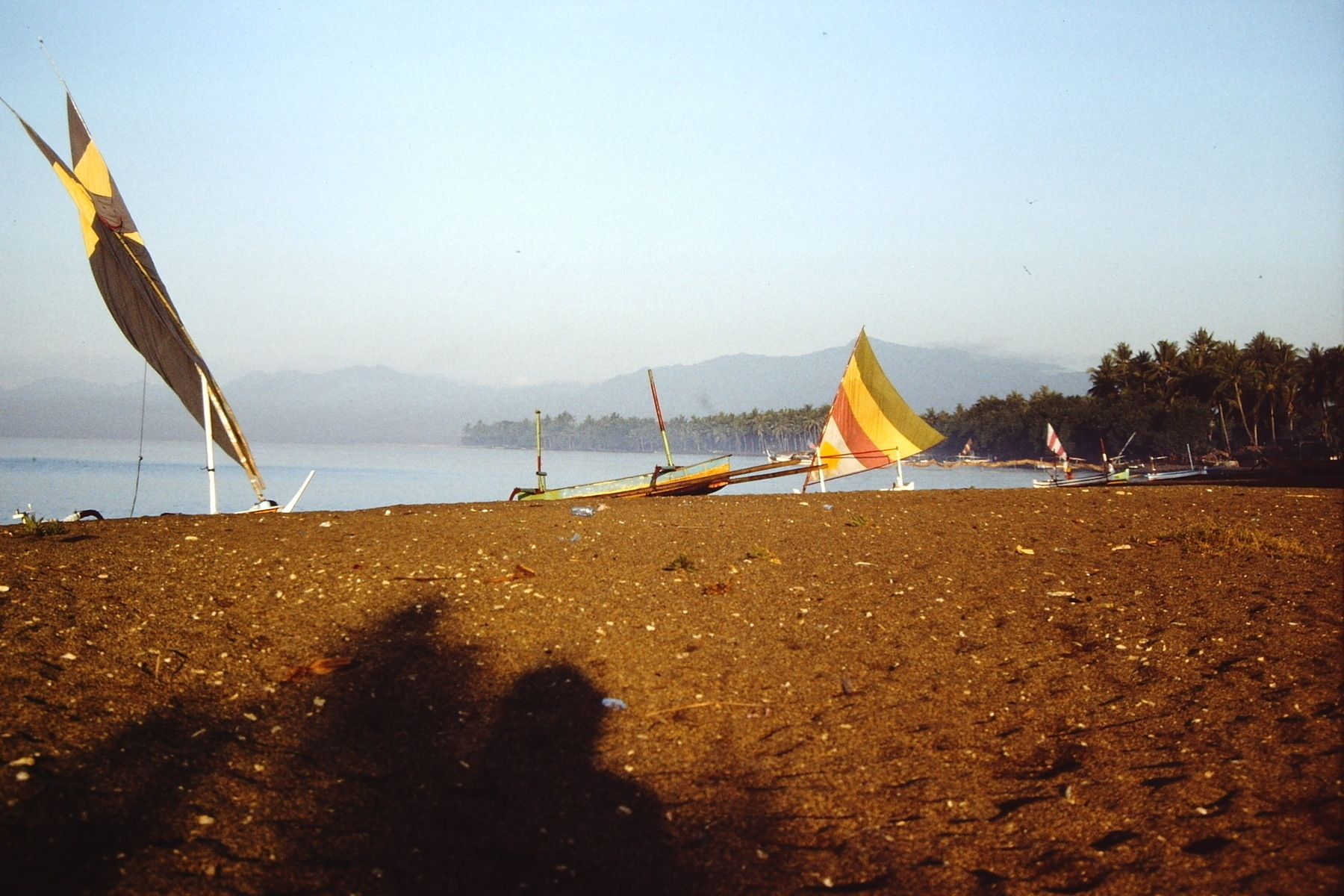 A tranquil beach scene features colorful sailboats on a sandy shore with calm waters and distant mountains in a haze. Probably taken at sunrise.