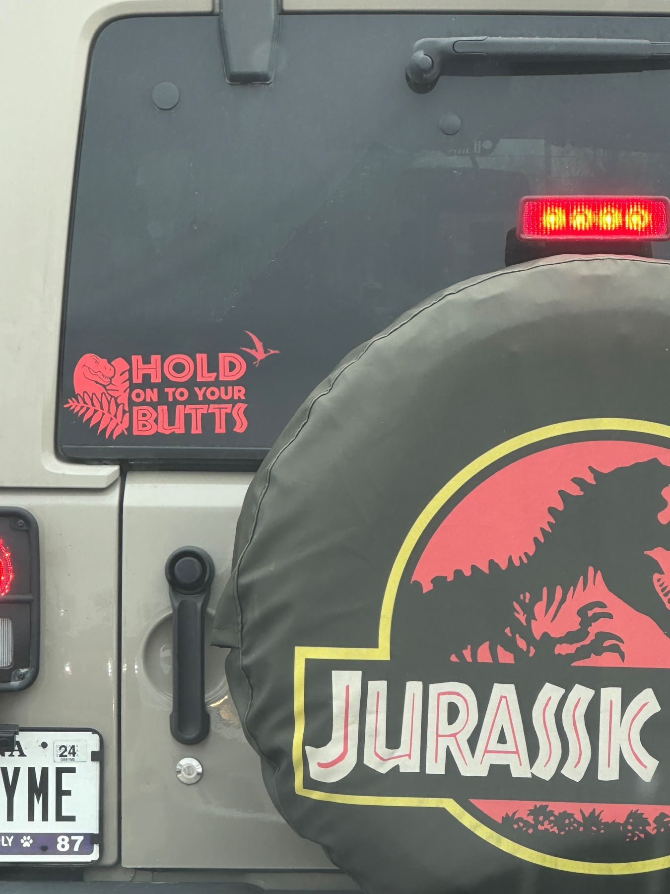 A vehicle’s rear spare tire cover featuring a stylized “Jurassic” logo with a dinosaur silhouette and the phrase “HOLD ON TO YOUR BUTTS” above it on the vehicle’s body. The car has a license plate at the bottom.