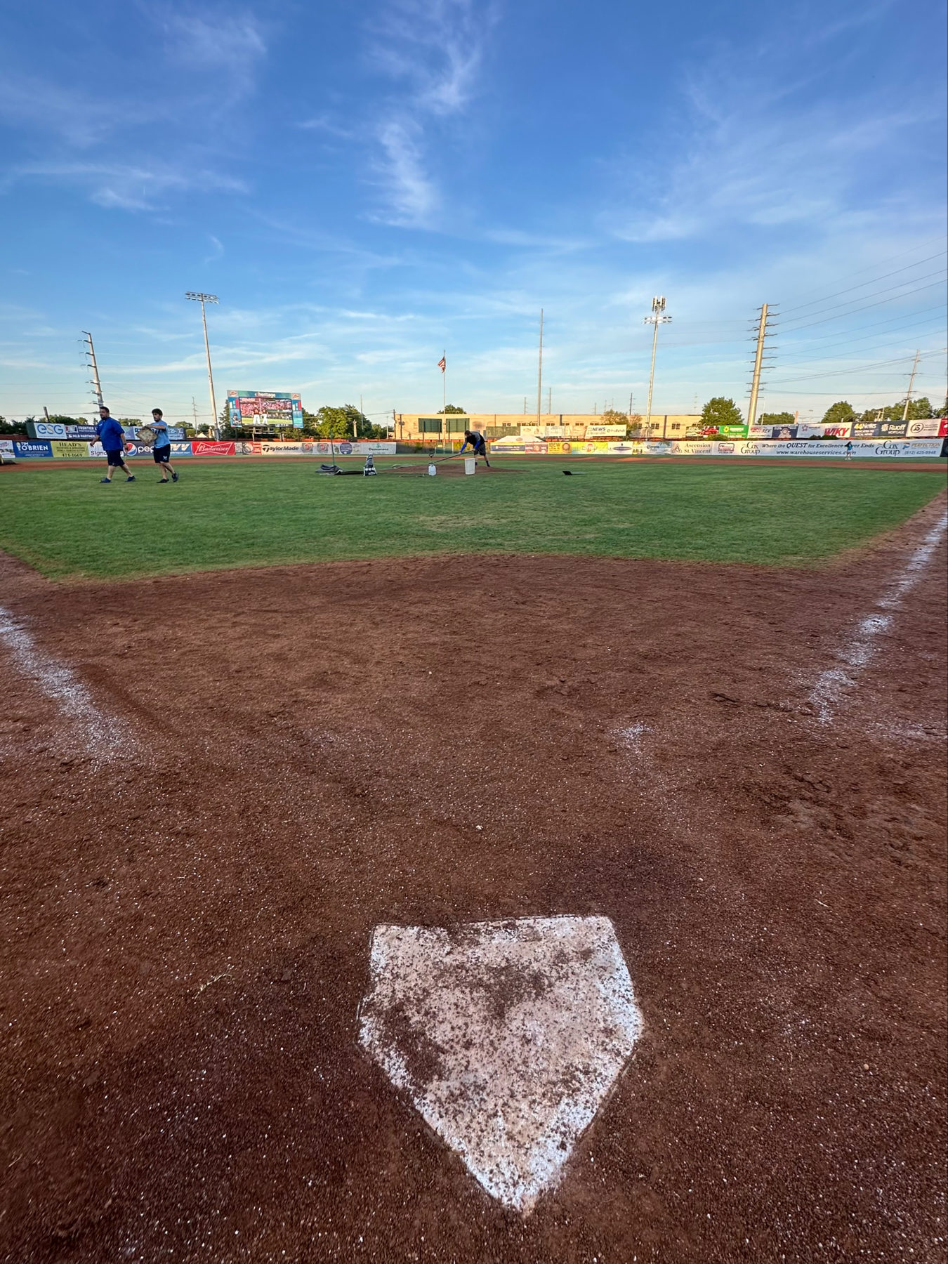A baseball field viewed from behind home plate. The infield dirt is prominent, and the green outfield stretches towards the horizon. Several players are seen near the pitcher’s mound, engaging in pre-game activities. The sky is clear with a few wis